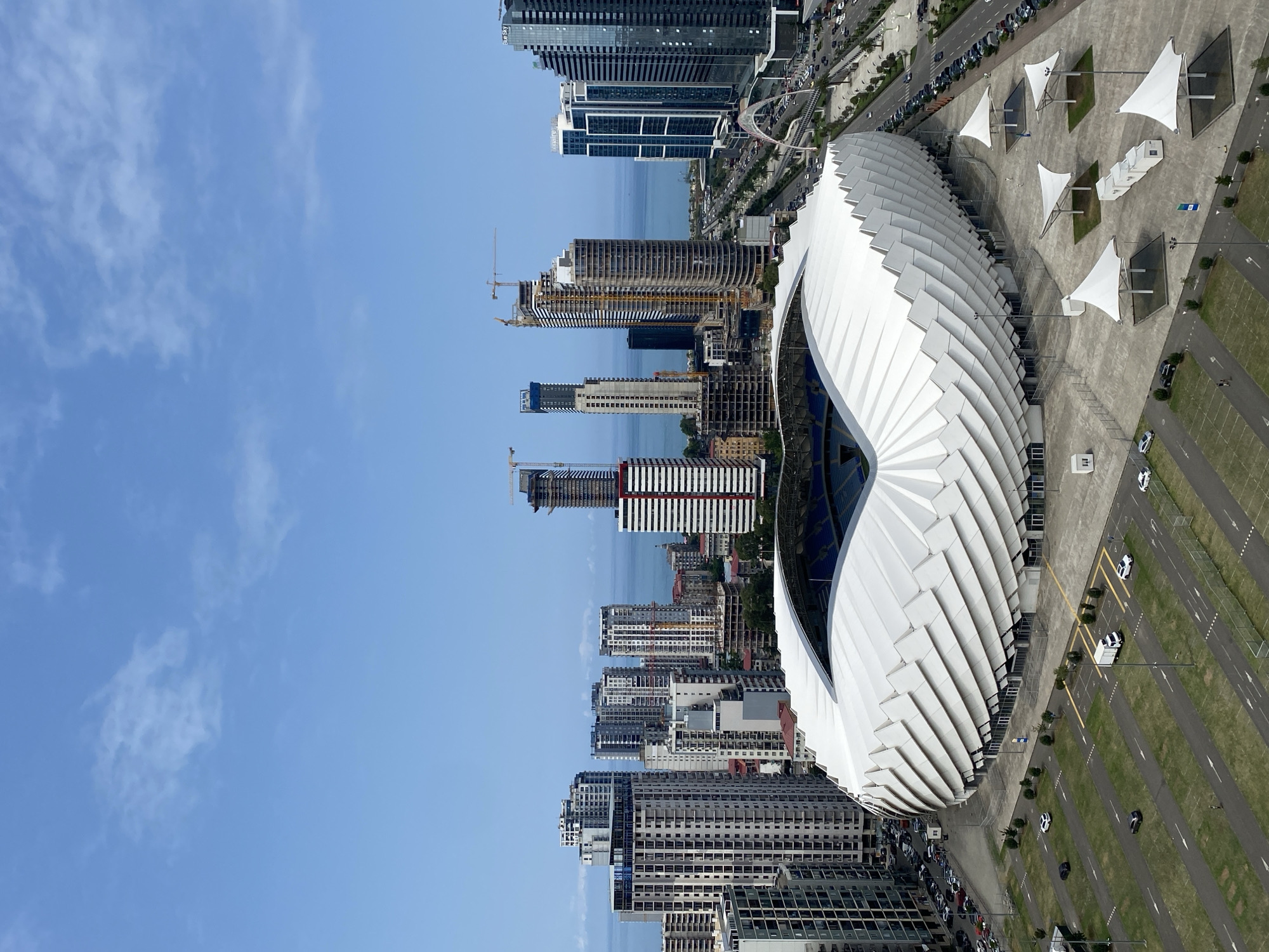The image captures a panoramic view of the Batumi Stadium, also known as the Adjarabet Arena, situated in the coastal city of Batumi, Georgia. The modern stadium, characterized by its unique, wave-like white roof structure, is the focal point of the scene. Surrounding the stadium is a vast paved area with parking lots, some featuring parked cars and a white delivery truck. Several white, tent-like canopies are erected on the plaza.
Beyond the stadium, the cityscape unfolds with numerous high-rise buildings, including modern residential and commercial towers, some of which are still under construction, evidenced by cranes visible on several structures. The Black Sea forms a distant backdrop, with a clear blue sky overhead dotted with scattered white clouds. A busy road with cars and a distinctive arched bridge can be seen to the right of the stadium, connecting it to the broader urban infrastructure. The overall scene depicts a contemporary urban environment with significant architectural development.