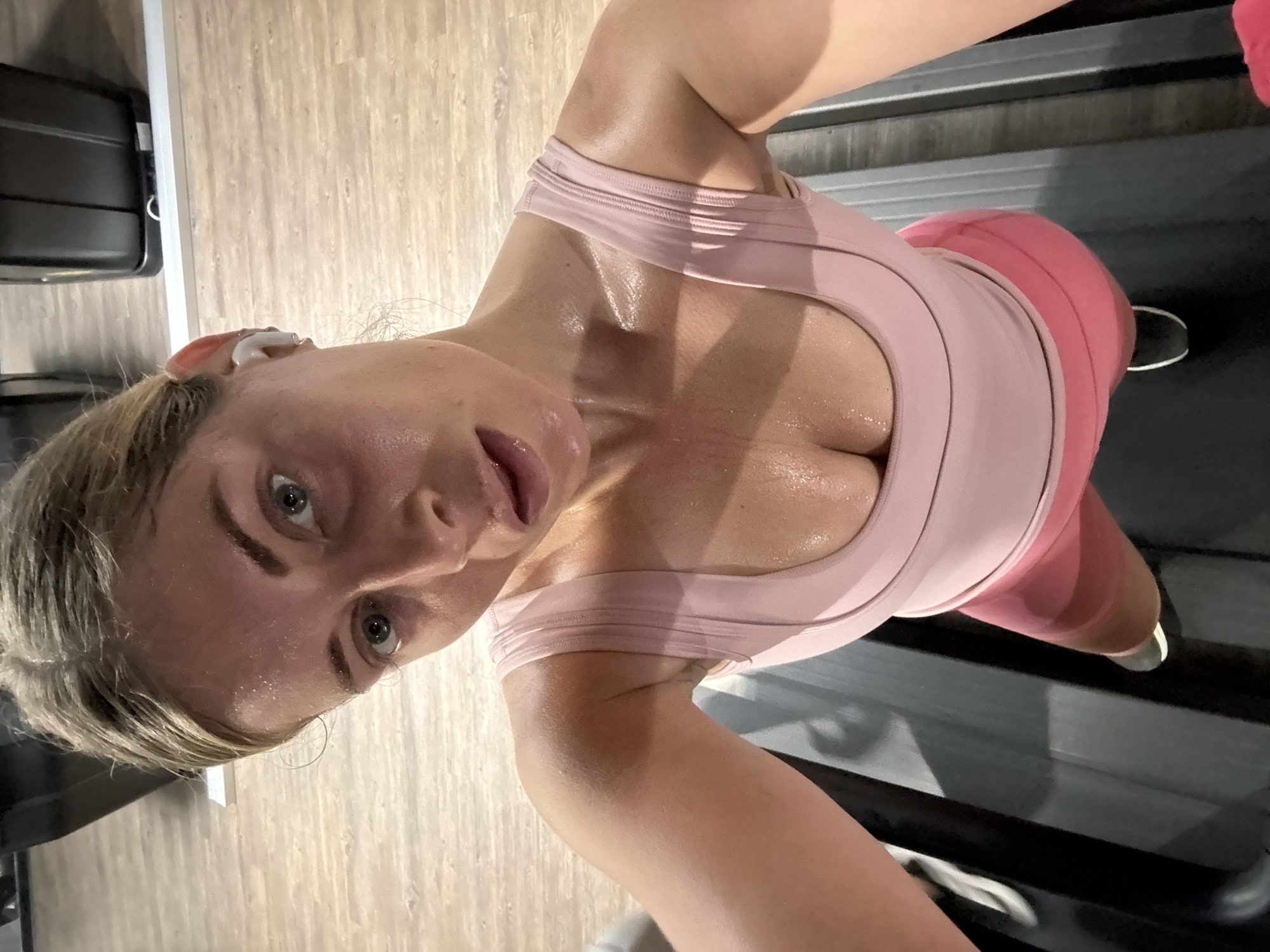 A woman is standing on a treadmill. She is wearing a pink workout top and pink leggings. She has blonde hair and is wearing an earbud. The woman is facing the camera and her face is sweaty. The treadmill is black and the floor is light brown.