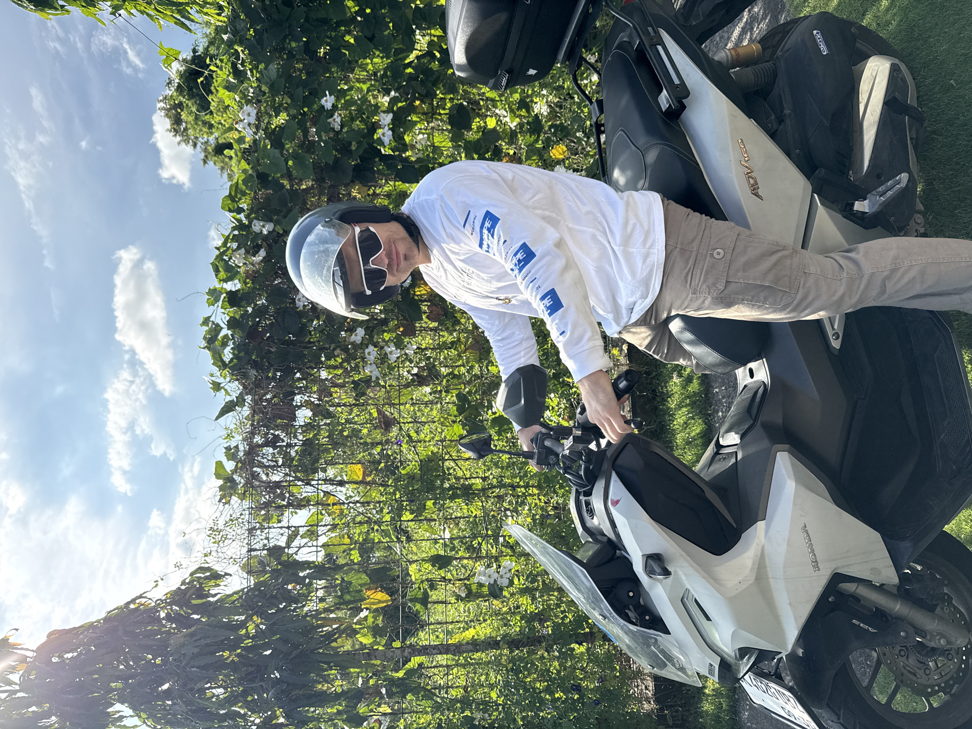 A person wearing a helmet and sunglasses is sitting on a white and gray Honda ADV 160 motorcycle. The motorcycle is parked on grass next to a fence covered in green vines and white flowers. The person is wearing a white long-sleeved shirt with blue text and khaki-colored pants. The motorcycle has a black cargo box mounted behind the seat. The location is Buduk, Indonesia.