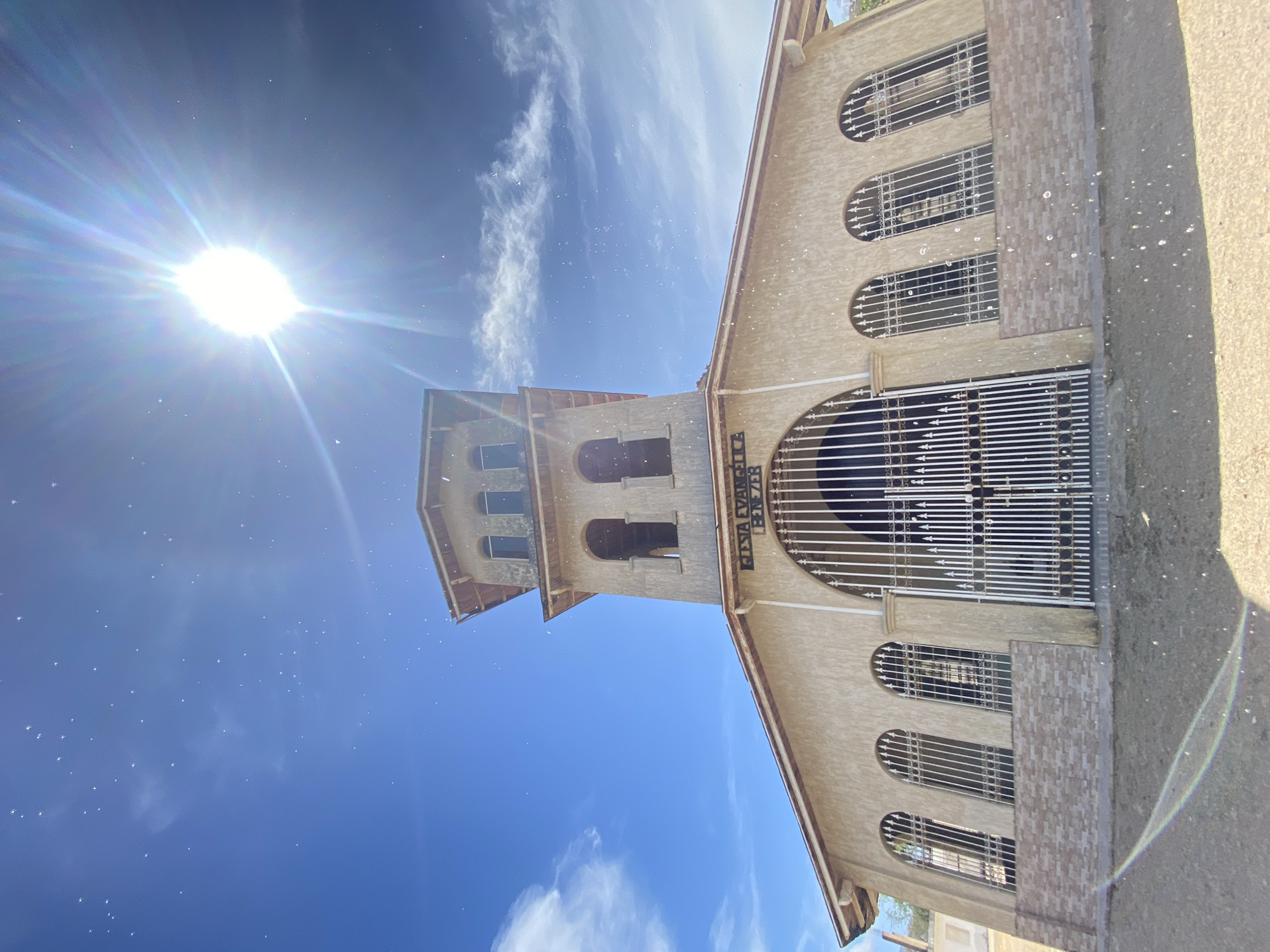 The image shows the Iglesia Evangélica Ebenezer (Ebenezer Evangelical Church) in Juan Griego, Venezuela. The building has a bell tower, arched windows with white bars, and a large arched gate. The sun is bright in the blue sky.