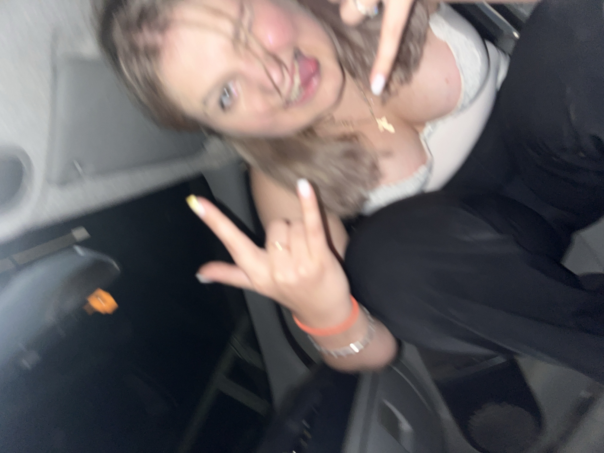 The image shows a young woman in a car. She has blonde hair and is wearing a white top and black pants. She is making a "rock on" gesture with her left hand and a peace sign with her right hand. She is sticking her tongue out and winking at the camera. There is a gold cross necklace around her neck. The background is dark, suggesting it is nighttime or inside a dark car. The car appears to be in motion. There is an orange object visible in the upper left corner of the image which could be a car accessory or part of the car's interior.
