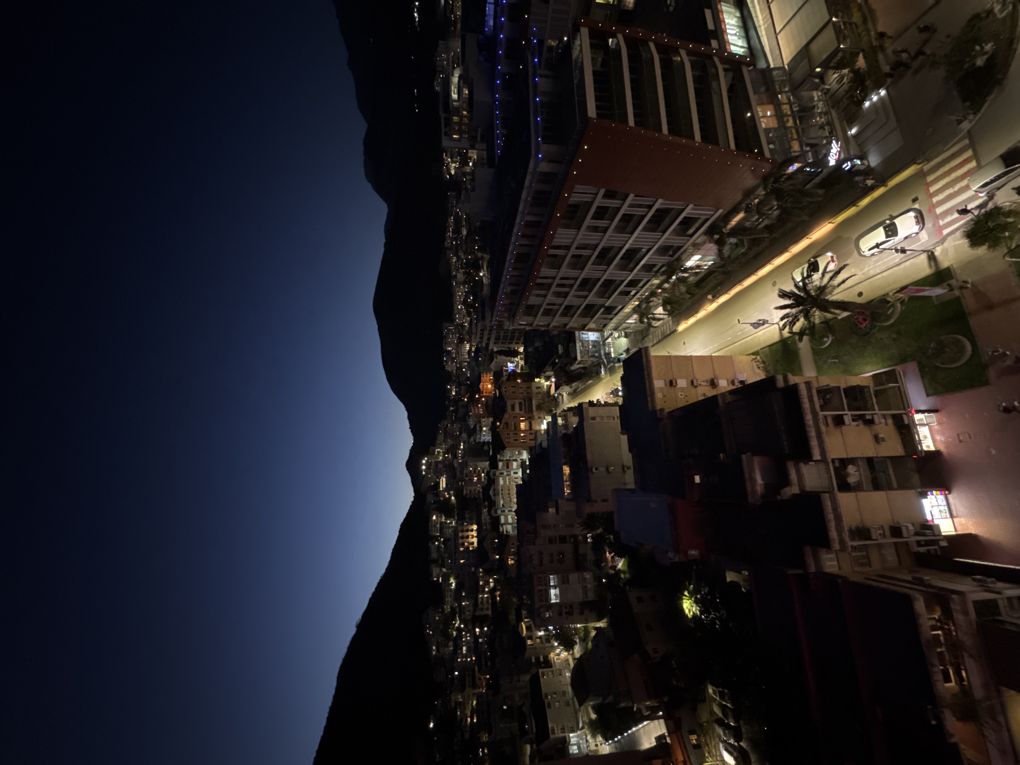 The image is an aerial view of a city at night, likely in Budva, Montenegro, looking toward the mountains. A dark mountain range is visible on the left, contrasting with the city lights. Buildings are densely packed, with numerous lights illuminating their windows and streets. A palm tree is visible on the side of the street. Cars are parked along the street. A tall building with visible balconies is situated in the right corner. The sky transitions from a dark blue to black above the mountains.
