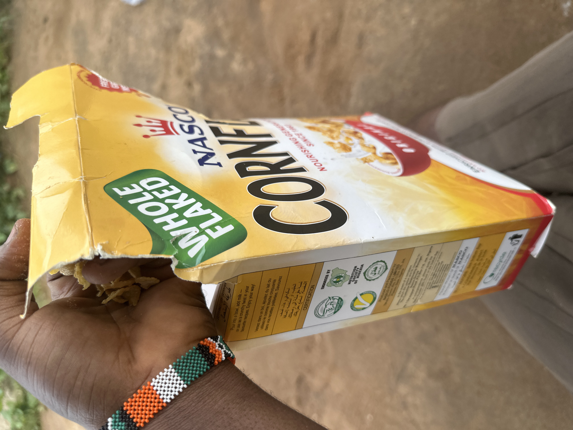 A person is holding a box of NASCO Corn Flakes, with the box open and some flakes visible. The box is yellow and white, and the text "WHOLE FLAKED" and "NASCO CORNE" is visible. The person has dark skin and is wearing a beaded bracelet. The location is not discernible to be Ikot Ekpene, Nigeria.