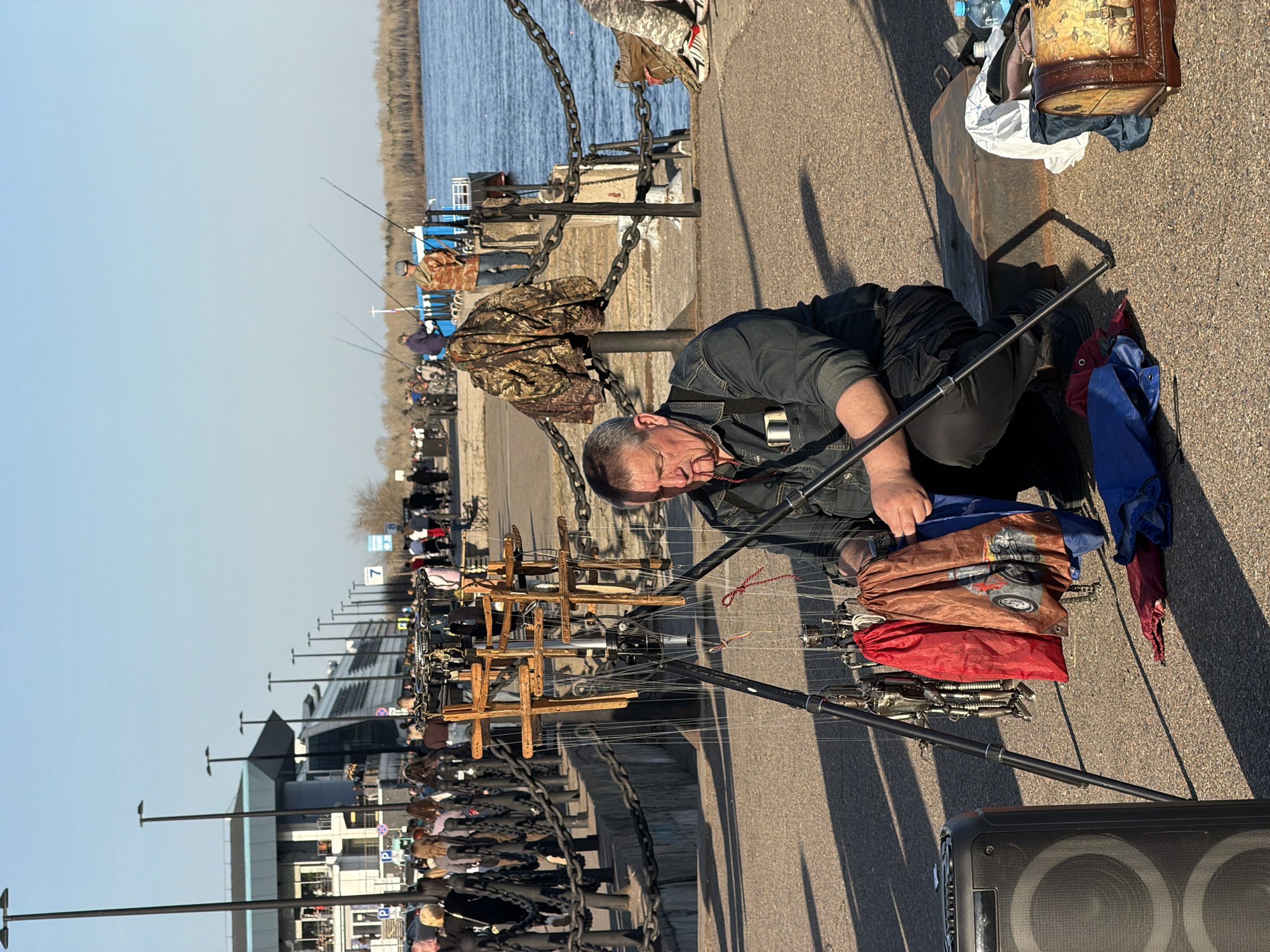 The image shows a man in Krasnoyarsk, Russia, participating in a traditional fishing event. He is squatting on a paved surface next to a river, with fishing rods extended over the water. The man is middle-aged and wearing a dark-colored denim shirt or jacket. He is focused on his fishing equipment, which is organized in bags near him. Other people are lined up along the riverbank, also fishing. A chain barrier runs along the edge of the pier or embankment. There is a building and signage visible in the background. A speaker is visible near the man.