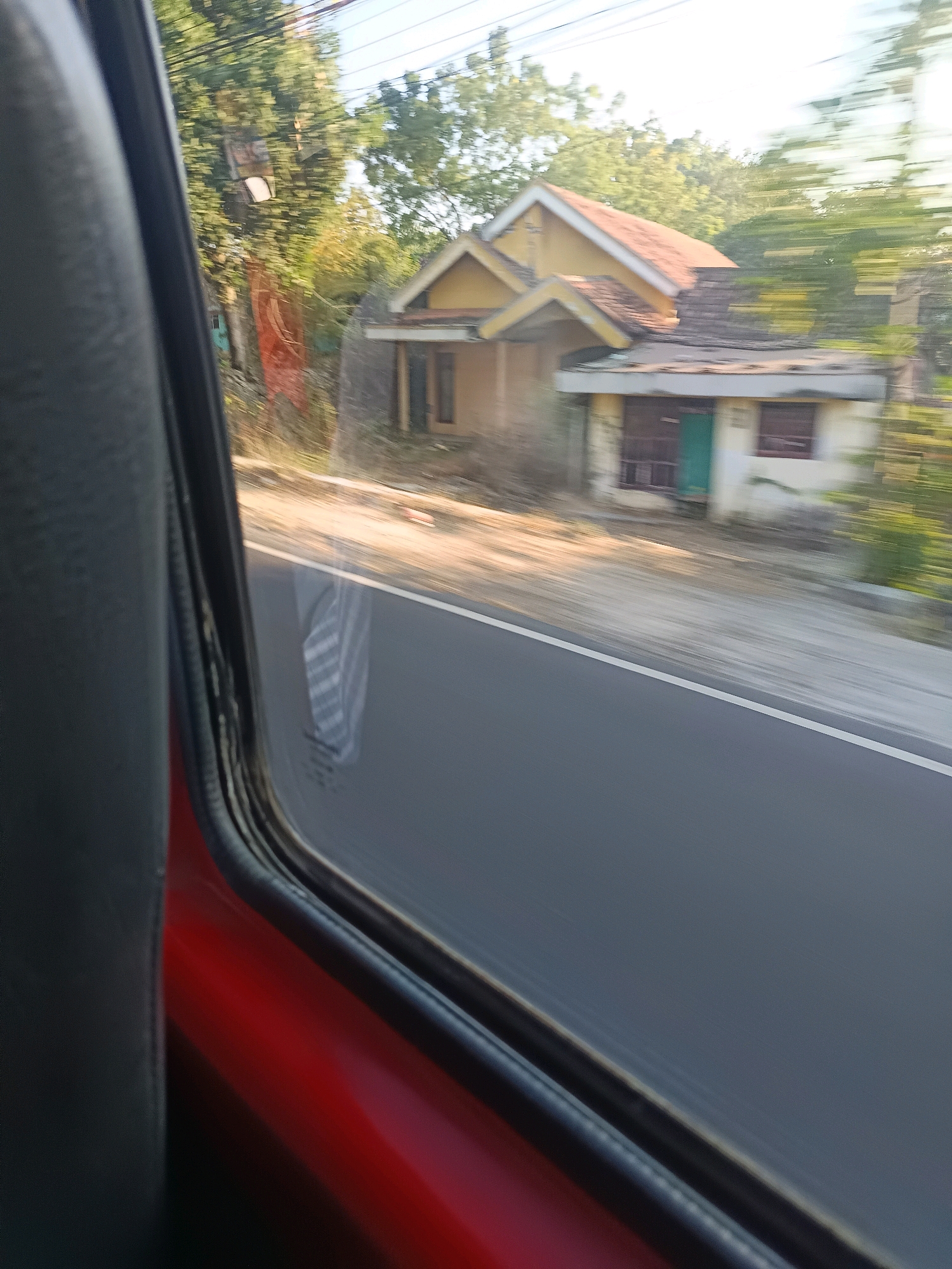 The image captures a view from inside a vehicle traveling on a road in Patarongan, Indonesia. The perspective is from the passenger seat, looking out the window. The foreground is dominated by the interior of the car, with a dark grey seat and a red car door visible on the left. The window frame is black.
Outside the window, a road with a white line marking the lane is visible, indicating movement. To the right of the road, there is a blurred scene of trees and two small, simple houses. The houses appear to be single-story structures with sloping roofs, one painted yellow and the other cream-colored. One house has a green door. The movement of the vehicle has caused motion blur in the outdoor elements, particularly the trees and the houses, making them indistinct. The sky is a clear, bright blue. The lighting suggests it is daytime, likely morning or afternoon.
