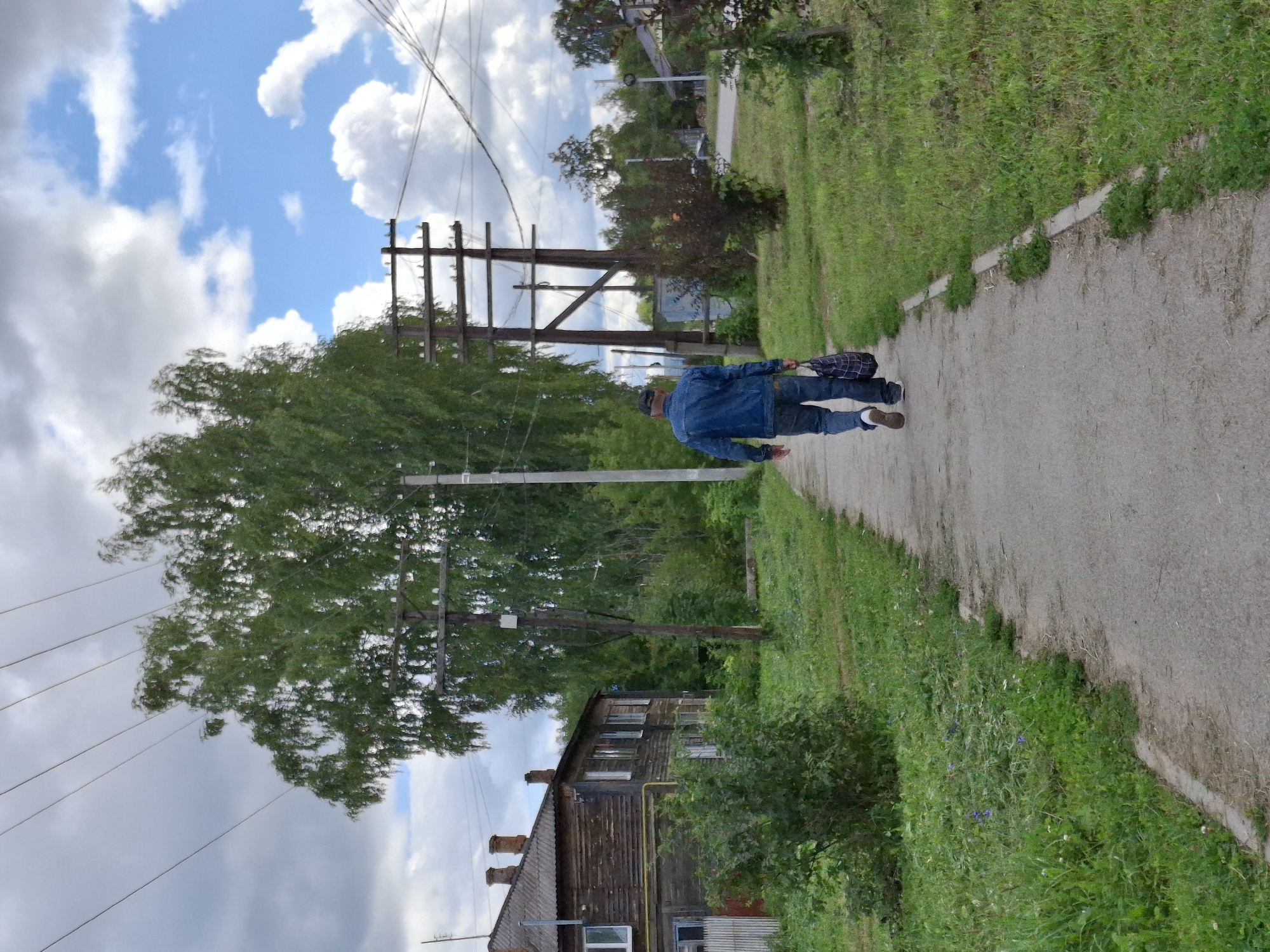A person walks away from the camera on a paved sidewalk in the city of Elovo, Russia. The individual is wearing a blue denim jacket, blue jeans, and white-soled shoes, and is carrying a patterned tote bag in their right hand. The sidewalk is bordered by a strip of grass and vegetation on the left, and a grassy area with scattered small plants and a sidewalk curb on the right. In the background to the left, a weathered, wooden two-story building with a grey shingled roof and visible chimneys stands amongst trees. To the right and behind the person, there are several wooden utility poles with visible wires stretching overhead. Further in the background, more trees and some residential structures are visible under a partly cloudy sky with blue patches. The overall scene depicts a street in a residential area of Elovo on a daytime.