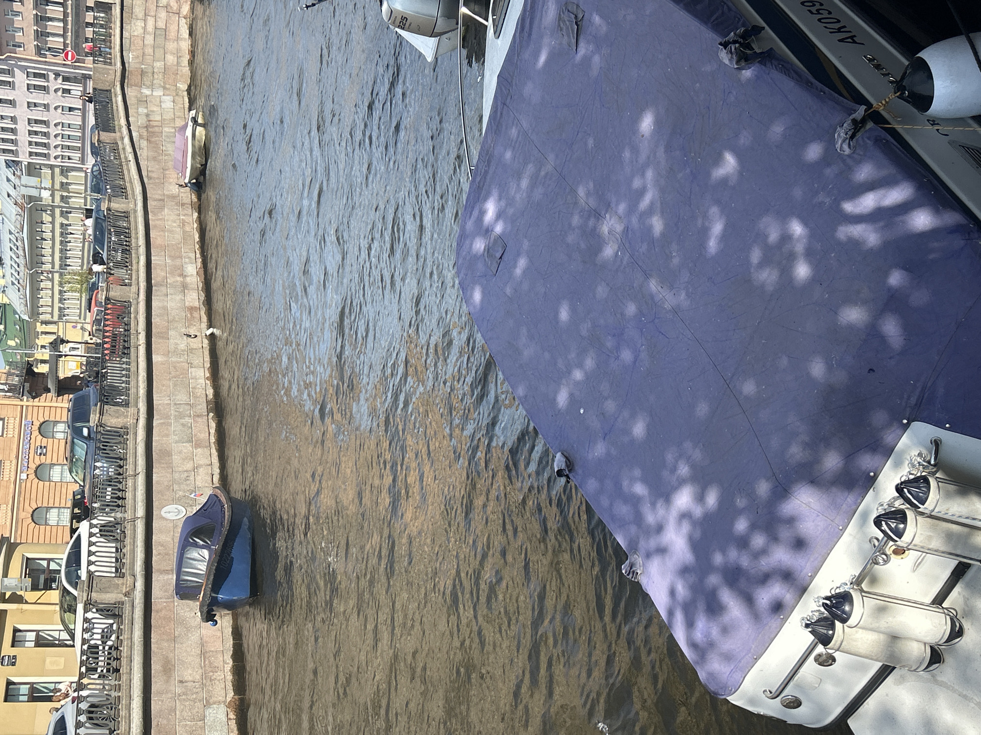 In an urban waterway, a blue motorboat with a covered cabin is moored near a stone embankment. Beyond the boat, a second boat with a light-colored cover is partially submerged. A larger boat, covered with a dark blue tarp, is in the foreground. Cars and pedestrians are visible on the street bordering the canal. The environment suggests a sunny day with dappled sunlight filtering through trees onto the water and boats.