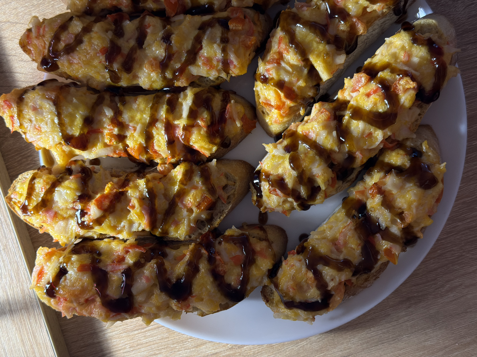 A close-up, overhead shot captures a white plate filled with several pieces of toasted bread topped with a creamy mixture and drizzled with a dark sauce. The bread appears to be baguette slices, toasted to a golden brown. The topping is a pale, golden-yellow mixture with visible chunks of reddish-orange, likely crab meat, and possibly some cheese. The dark sauce, which resembles teriyaki or balsamic glaze, is artfully drizzled in a crisscross pattern over the topping.
The food is arranged on a white, circular plate. The plate itself sits on a light brown wooden surface, possibly a table. Sunlight casts shadows across the plate and the food, indicating it might be daytime, with the sun coming from the left side of the frame. There are no people visible in the image. The setting appears to be indoors, in a casual environment. The focus is solely on the food, highlighting its texture and presentation. No text is visible in the image.