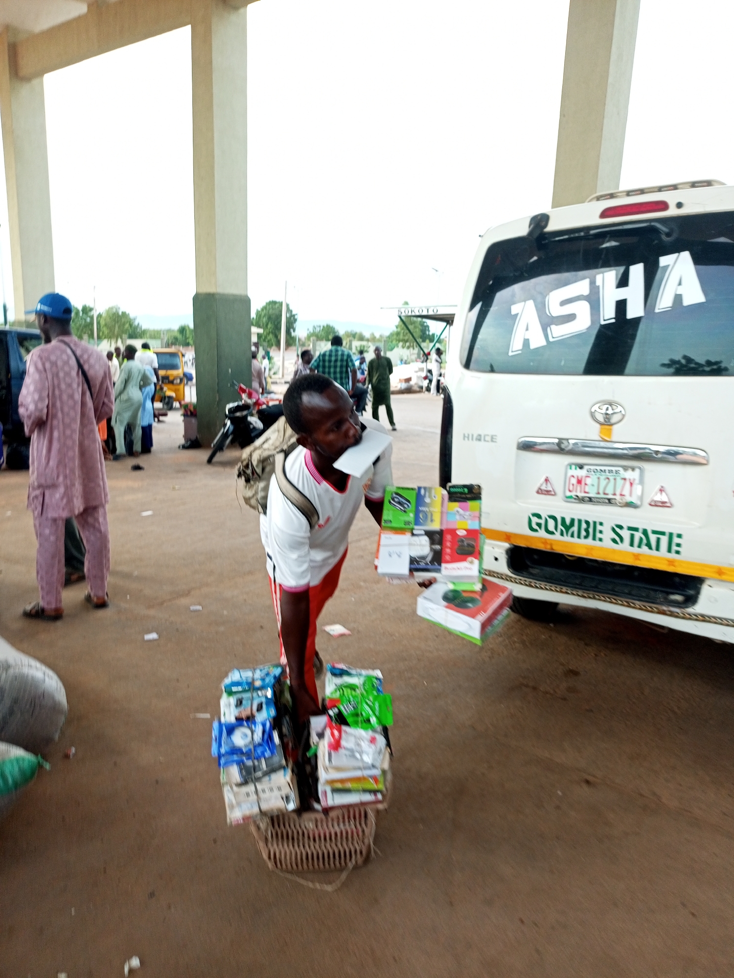 The image depicts an outdoor scene, likely a transportation hub or market area, in Gombe State, Nigeria. In the foreground, a man with dark skin, wearing a white t-shirt with red trim and red trousers, is holding a piece of paper to his mouth. He is surrounded by stacks of small boxes, seemingly electronics or accessories, placed on a woven basket and on the ground. Behind him, a white van with the inscription "ASHA" and a license plate reading "GME-121ZY" is parked. The van also has "GOMBE STATE" written on its rear. Other people are visible in the background, some walking, and a yellow motorized tricycle is present. The environment appears to be a dusty, open area with concrete flooring, bordered by covered structures with large columns and some trees in the distance. The overall lighting suggests daylight.