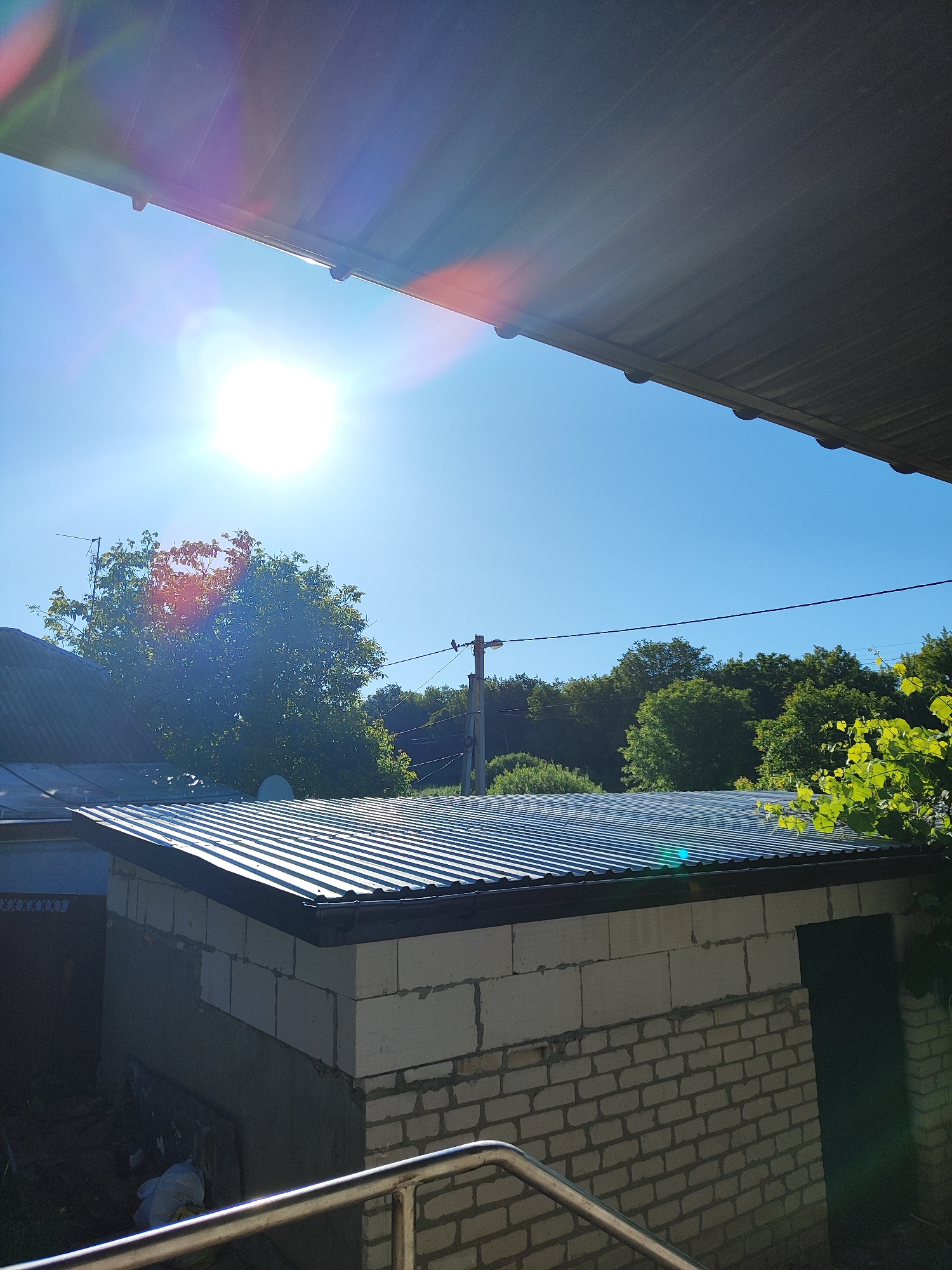 A bright, sunny day is depicted in this outdoor scene. The sky is a clear, vibrant blue, with the sun prominently shining and creating lens flare. In the foreground, a corrugated metal roof extends from the top edge of the frame, casting a shadow downwards. Below this, another corrugated metal roof covers a rectangular structure built with white cinder blocks and some white bricks. The walls of the structure are partially constructed and incomplete, with some visible mortar. The structure has a dark doorway on its right side. In the background, lush green trees form a dense line against the horizon. A utility pole with wires stretches across the middle ground, and a bird is perched on one of the wires. To the left, part of a dark-colored roof of another building is visible.