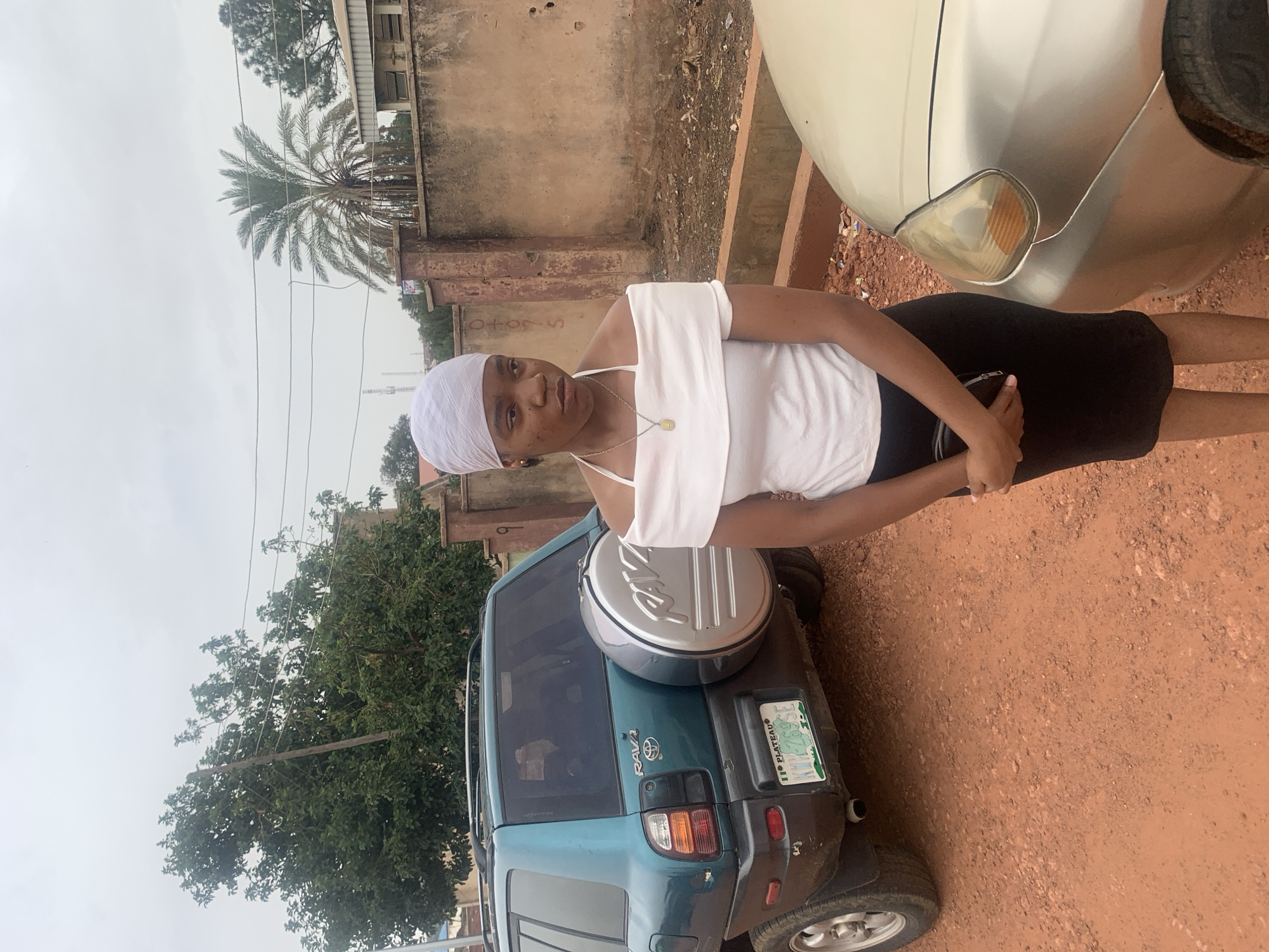 A young woman stands outdoors next to a teal-colored RAV4. She wears a white top, a black skirt, and a white headscarf. Part of a light-colored car is visible on the right. A wall and buildings are in the background, as well as trees. The ground is red dirt. This image could be of Jos, Nigeria based on car registration on the RAV4.