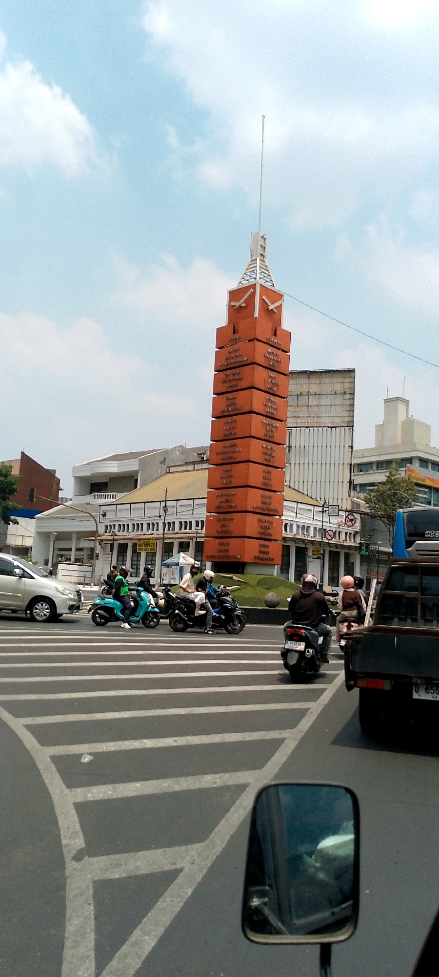 The image captures a busy street scene in Bandung City, Indonesia, likely near the iconic Monumen perjuangan rakyat jabar. A tall, orange, brick-like monument with text on its surfaces dominates the center of the frame, reaching towards a bright, partly cloudy sky. In the foreground, a pedestrian crossing with white stripes marks the intersection. Several motorbikes are in motion, navigating the road. One motorbike, ridden by a person in a green jacket, is in the left lane of the crossing. Two other motorbikes follow, one with two passengers, all heading in the same direction. Further down the road, a pickup truck is visible, and a silver car is in the left lane. The background reveals various buildings, including a white building with a patterned facade and a tall, modern structure behind the monument. The overall impression is that of a typical day in a bustling urban environment.
