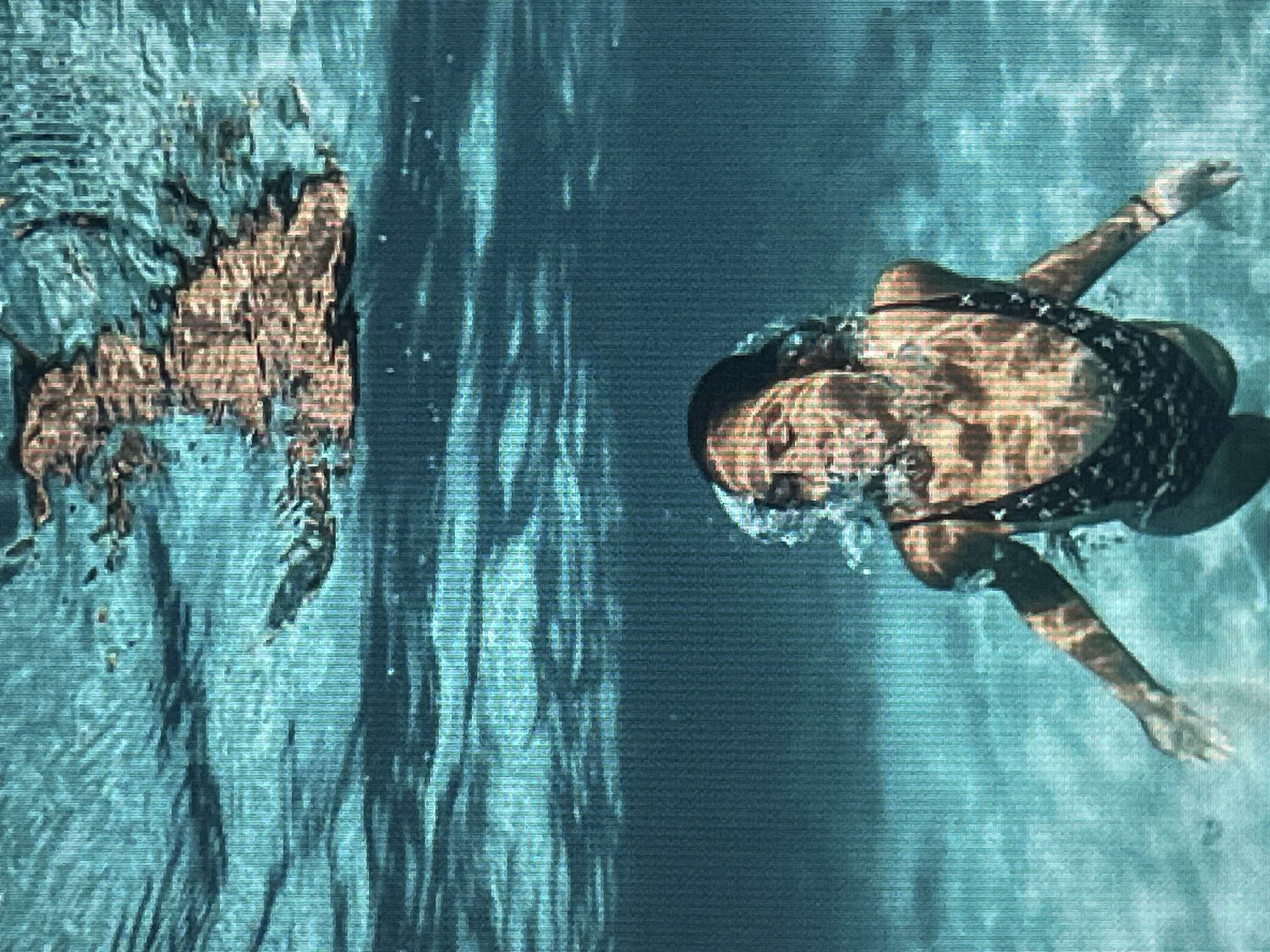 This is a horizontal, underwater shot of two people swimming in a pool. On the left is a person with fair skin, wearing a tan-colored swimsuit. They are positioned vertically, with their head down and legs up, their body forming an "L" shape. On the right, a person with darker skin is swimming on their back. They are wearing a black swimsuit with a white speckled pattern. Their head is to the left of the frame, and their body is angled diagonally towards the bottom right. Their left arm is extended in front of them, reaching forward, while their right arm is bent at the elbow. The water is a teal color, with light reflecting off the surface in wavy patterns. There is no visible text.