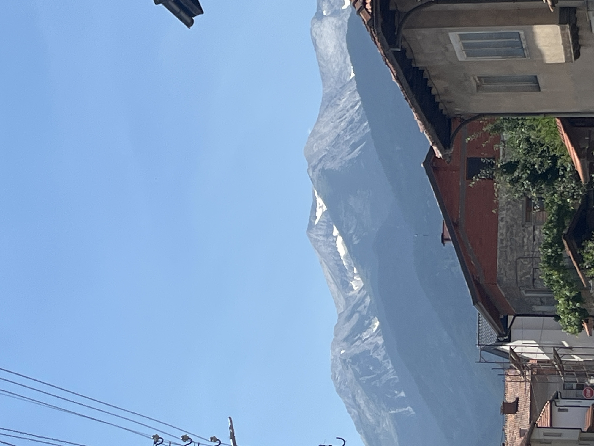 The image shows a mountainous landscape behind buildings in Bansko, Bulgaria. The sky is clear and blue. The mountains in the background are tall and rugged, with patches of snow visible on their peaks. In the foreground, there are several buildings with red tile roofs and stone facades. Some of the buildings appear to be older, while others show signs of recent construction or renovation, with scaffolding visible on one structure. Electrical wires are visible in the upper left corner of the image, strung across the sky. The overall impression is a blend of natural beauty and urban development in a mountainous region.