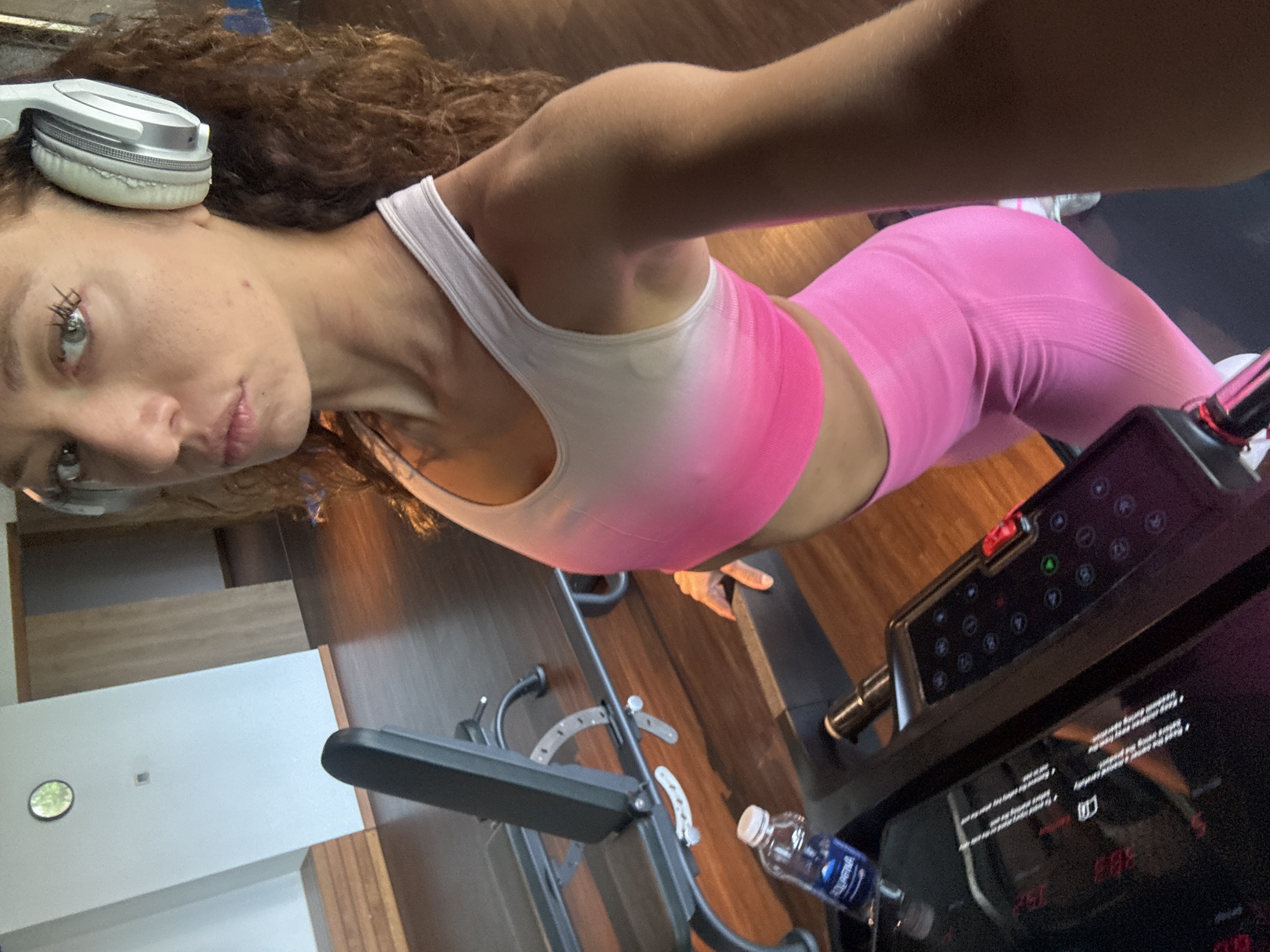 A woman with brown curly hair wearing white headphones, a white and pink sports bra, and pink leggings stands on a treadmill. She is looking at the camera. A water bottle sits in a holder on the treadmill. The treadmill has a black console with red and green buttons. In the background, there are wooden floors and white cabinets. The location is unknown.