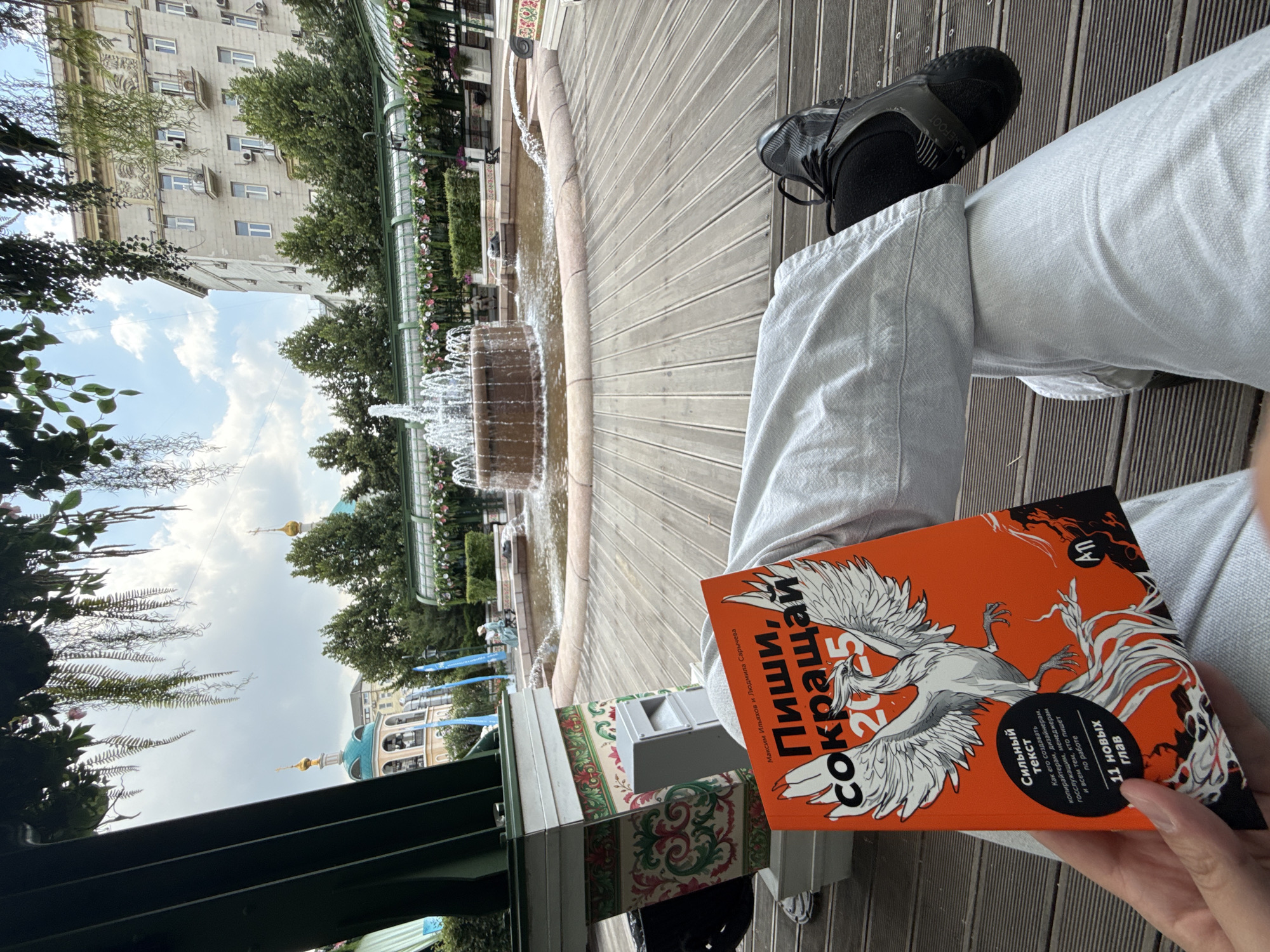 A person is sitting on a wooden deck, holding an orange book with a white phoenix on the cover. The book's title is "Пиши, сокращай 2025". The person is wearing light-colored pants and black athletic shoes. In the background, there is a fountain with water jets, surrounded by a paved area and greenery. Beyond the fountain, there are buildings with architectural details, including domes of a church, and trees. The sky is cloudy. The scene appears to be an outdoor public space, possibly a park or a cafe courtyard in Moscow, Russia, given the architectural style and the Cyrillic text on the book.