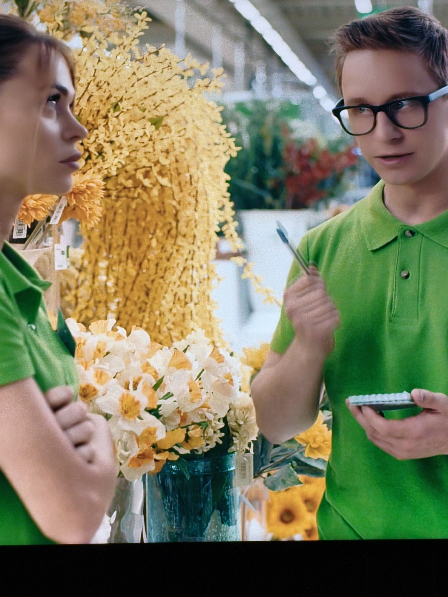 Two individuals, likely employees of a flower shop, are depicted in a brightly lit indoor setting, possibly a supermarket or a large florist. On the left, a woman with light brown hair and bangs is seen in profile, looking upwards and to her left. She is wearing a green collared shirt, with a portion of her arm visible. To her right, a young man with dark brown hair and black-rimmed glasses is looking towards the woman, holding a pen in his right hand and a small notepad or data entry device in his left. He is also wearing a green collared shirt. Behind them, an abundance of yellow flowers hangs in large arrangements, and in the foreground, a vase holds a bouquet of white and yellow daffodils. The background suggests an aisle with shelving and other floral displays. The location is not identifiable as Vladivostok, Russia, or any other specific city.