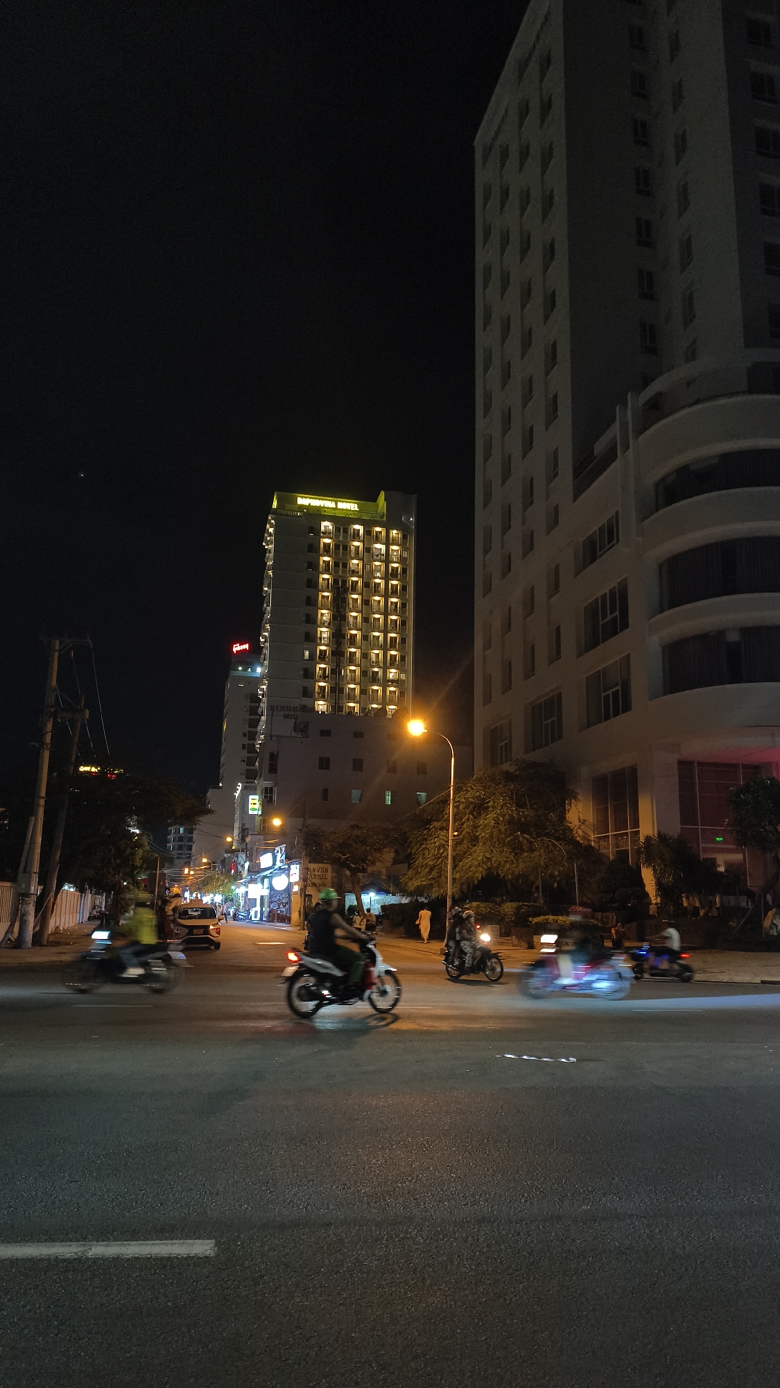 This is a night-time, eye-level, medium shot of a street intersection in Nha Trang, Vietnam. The scene is dominated by two tall buildings on the right and a slightly shorter, illuminated building in the background. The building on the far right is a modern, white high-rise with visible windows and curved architectural elements. To its left is another tall, white building with a more uniform facade. In the mid-ground, a brightly lit hotel, identified by a yellow sign at its top that reads "IMPERIAL HOTEL", stands out against the dark sky. Several floors of this hotel are illuminated, with rows of balconies visible.
The foreground shows a paved street with white lane markings. Several motorcycles are in motion, captured with motion blur. One motorcycle in the center foreground is white with red accents, ridden by a person wearing a dark helmet and a green shirt. To its left, another motorcycle is blurred with a rider in yellow. To the right, further motorcycles are also in motion, some with illuminated headlights.
Streetlights are visible, casting an orange glow on the road and surrounding areas. Trees with dark foliage are present along the sides of the street. The sky is dark, with only a faint hint of a distant celestial body visible in the upper left quadrant. The overall impression is of a busy urban street scene at night, characterized by vehicle traffic and illuminated urban architecture.
