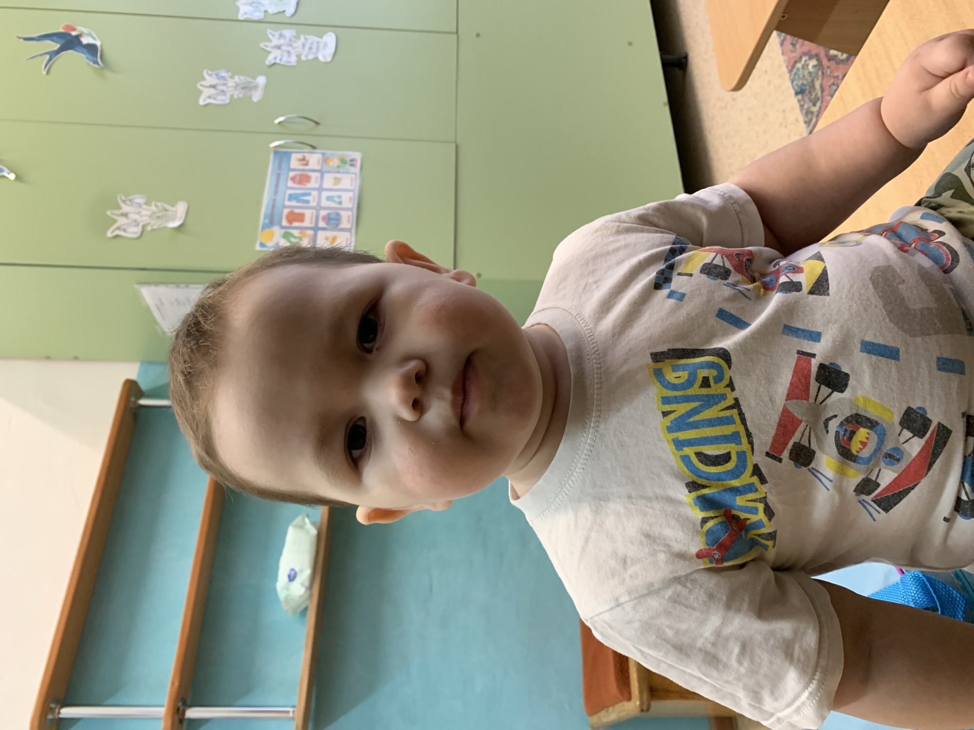 A young child is centered in the frame. He has light skin, dark eyes, and short brown hair. His cheeks are slightly flushed. He is wearing a white t-shirt with a colorful racing-themed print. The "RACING" print is visible in large, stylized blue and yellow letters.
The background includes a green cabinet with decorations and educational posters, a light blue wall with a wooden shelf, and a wooden chair with a patterned cushion.
The location is presumed to be Фоки, Russia, based on the image description request.