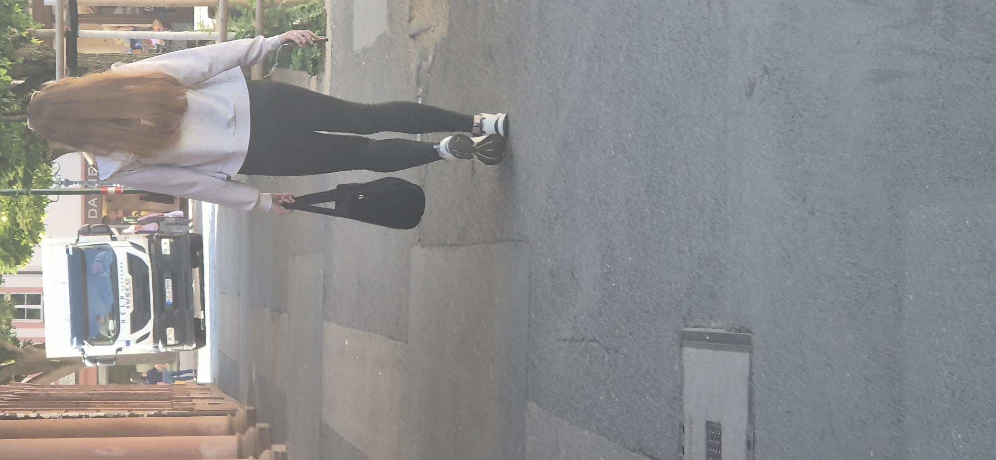 An image shows a person walking away from the camera down a street in Mainz, Germany. The person has long, red hair and is wearing a light-colored jacket, black leggings, and white athletic shoes. They are carrying a black tote bag in their right hand and have earbuds in their ears. In the background, a white Iveco truck is driving towards the camera, with two people visible in the driver's cab. The street is paved with grey stones and asphalt, with buildings on either side. The architecture suggests a European city, and the presence of the Iveco truck with German license plates confirms the location as Germany, likely Mainz. The lighting indicates it is daytime, with sunlight casting shadows on the ground.