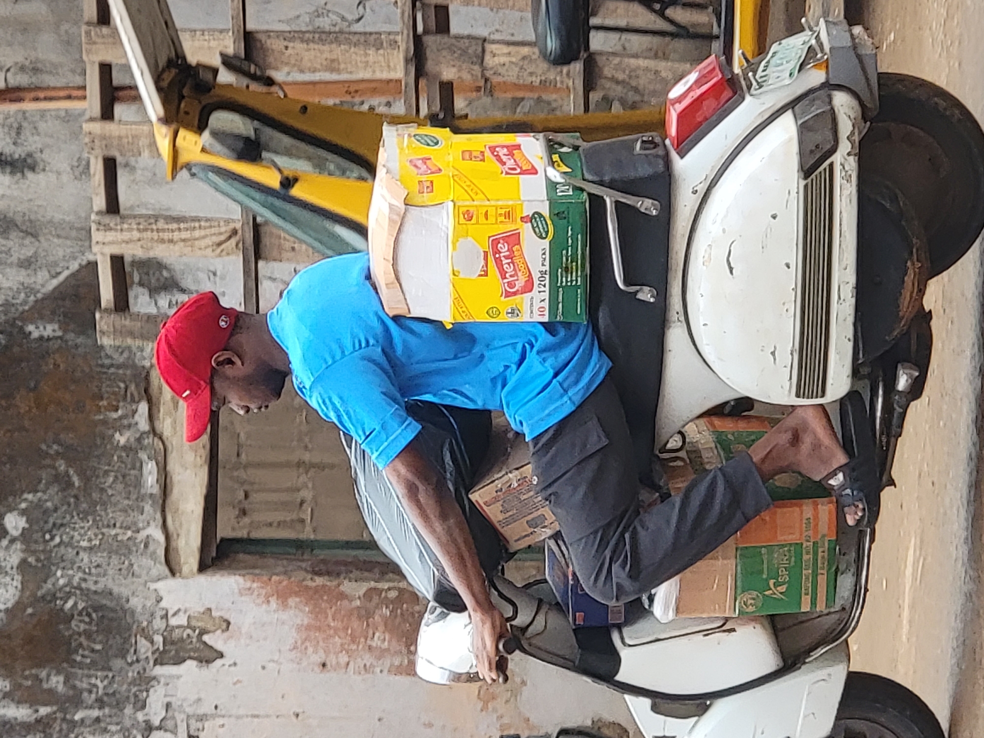 A man wearing a red cap and a blue t-shirt is seated on a white moped, seemingly in Maiburiji, Nigeria. He is surrounded by boxes of goods loaded on the moped. He is wearing black pants and sandals. The background shows a building in disrepair.