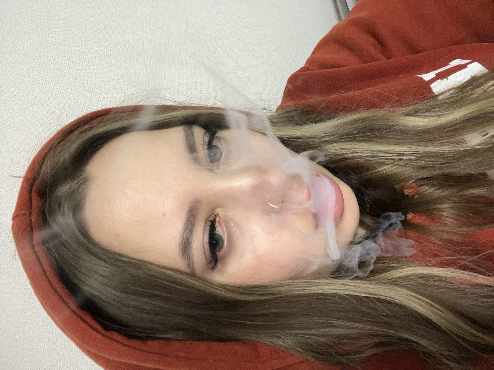 A woman with long, light brown hair wearing a red hooded sweatshirt is exhaling smoke from her mouth. The photograph appears to have been taken indoors against a light-colored wall. The location is not discernible from the visual information.