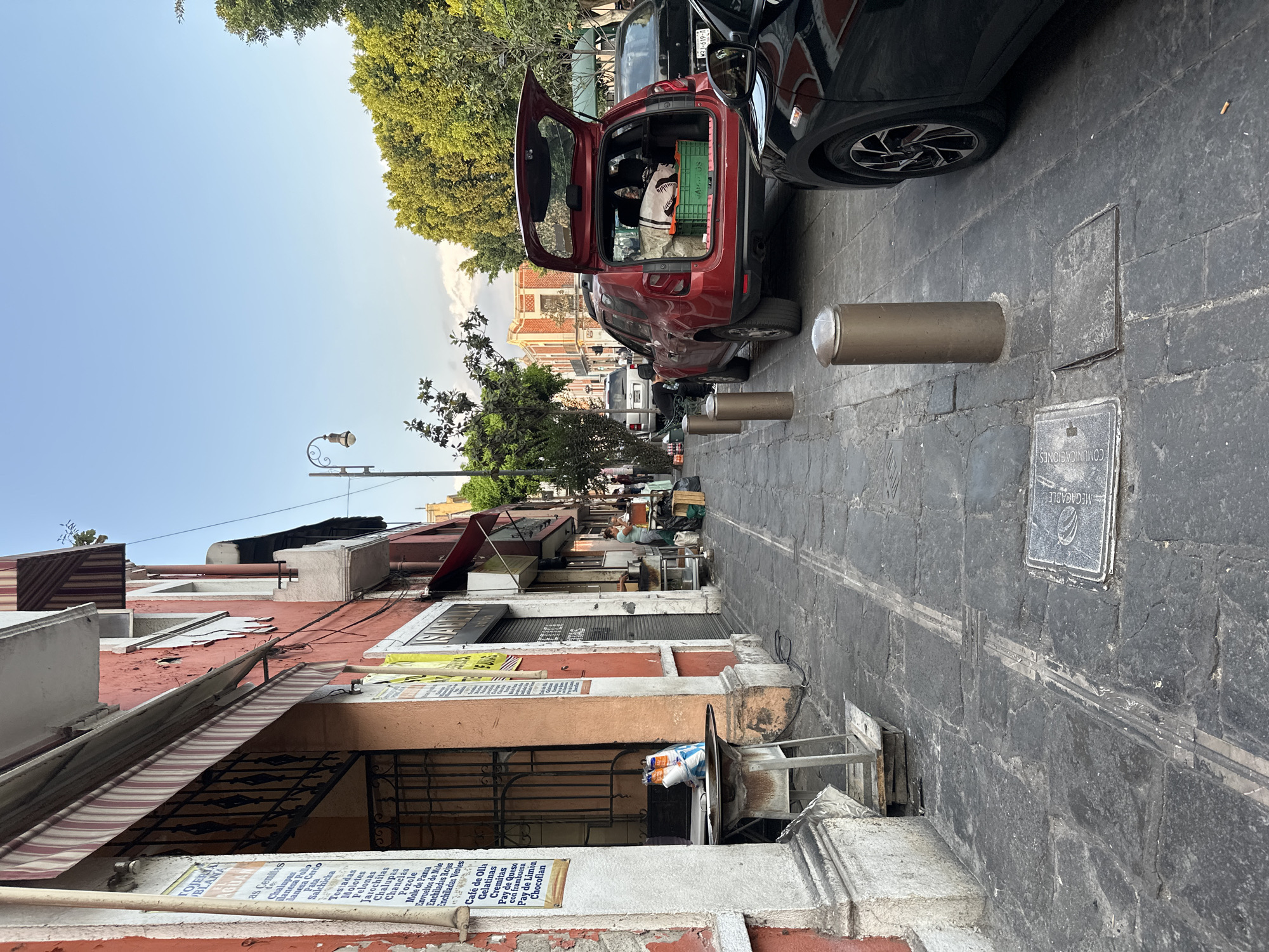 The image depicts a street scene in an urban environment. The perspective is street-level, oriented along the length of the street. Buildings line the left side of the image, with signs, awnings, and architectural details. One sign has text in Spanish about food. On the right side of the street, there are cars parked close to one another. One car has an open trunk revealing items inside. In between the vehicles are metal bollards. The street surface is made of cobblestones or similarly shaped pavers. Trees can be seen in the background. The sky is visible above the buildings, with a light blue hue. Overhead, utility wires are strung between buildings. The overall impression is of a busy street in a densely built area.