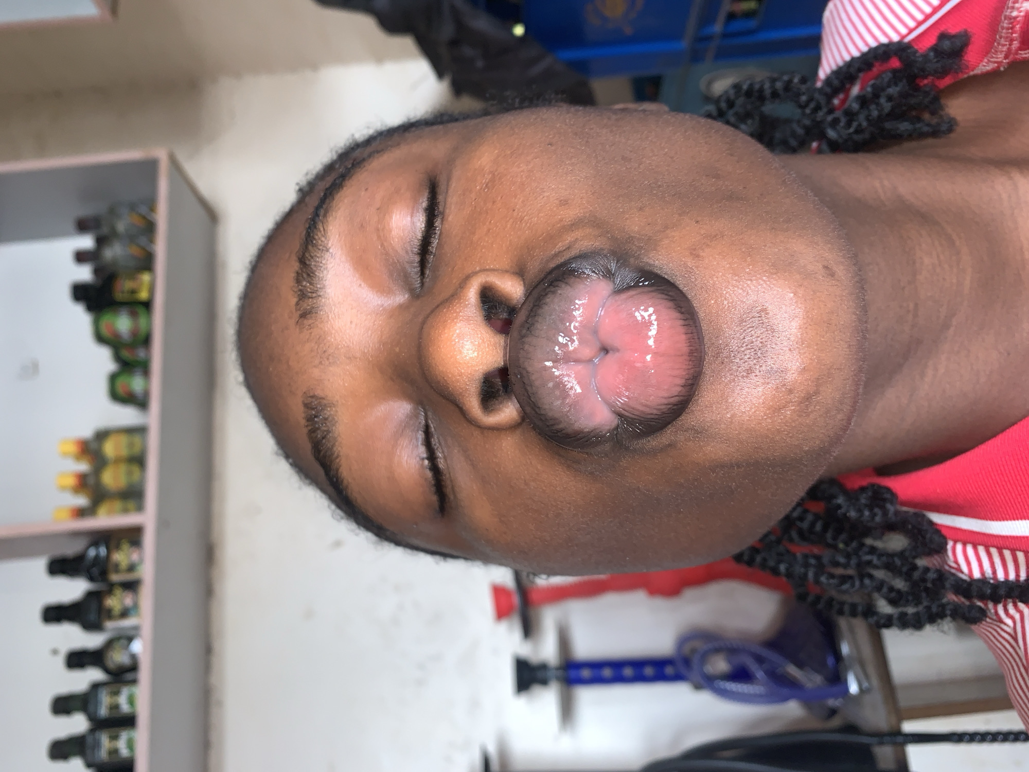 The image features a close-up portrait of a person with dark skin and braided hair, captured indoors in New Stadium, Nigeria. The individual's eyes are closed, and their lips are puckered outward in a distinctive, exaggerated expression, likely a playful pose.
In the background on the left, a light-colored shelving unit is visible, holding several bottles. These bottles appear to be various beverages, possibly spirits or wines, though their labels are not clearly legible. The person is wearing a red and white striped garment, which is visible on the right side of the frame. Towards the bottom right, parts of a blue object, possibly a hookah or a stand, can be seen. The walls in the background are a light, neutral color. The scene suggests a casual, indoor setting, possibly a home or a social gathering space, where a lighthearted moment is being captured.