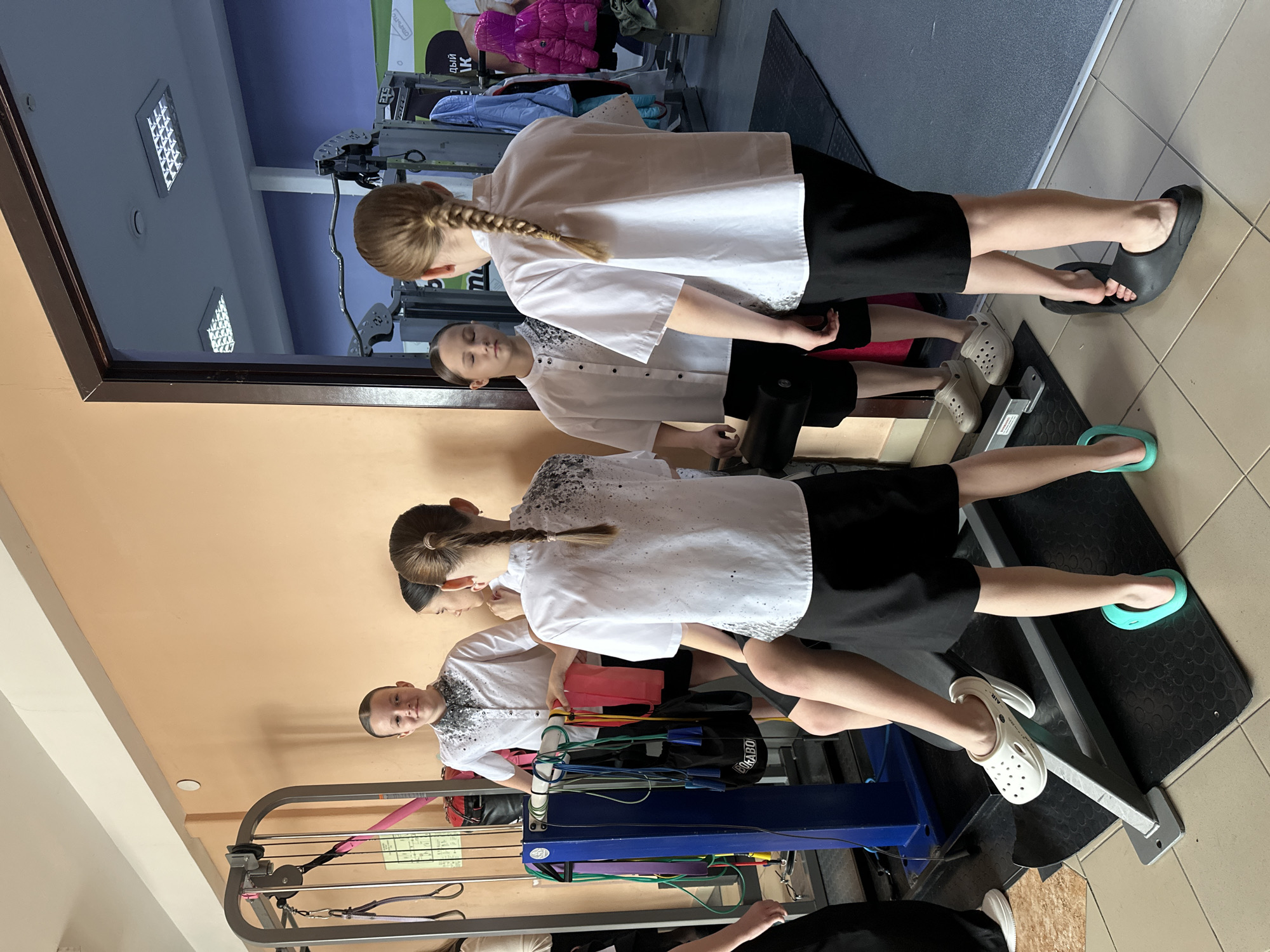 A photograph depicts three individuals standing in what appears to be a medical or physical therapy setting. The individuals appear to be two young girls and a young boy, all similarly dressed in white shirts and dark bottoms. The setting includes medical equipment and a reflective surface, indicating a clinic. It is unknown if the photo was taken in Votkinsk, Russia.
