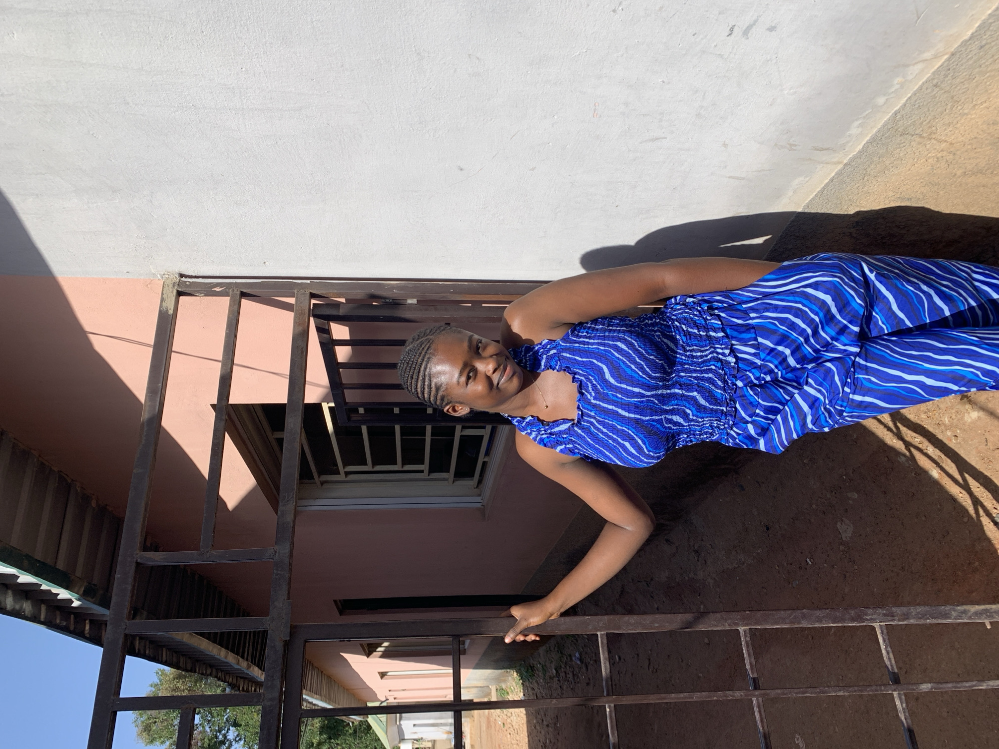 A woman stands outside a building in New Stadium, Nigeria. She is wearing a blue and white striped dress. She stands behind a dark metal fence. A shadow is cast to her right. The building behind her is two-toned, light grey and pink. There is also a window behind the fence.