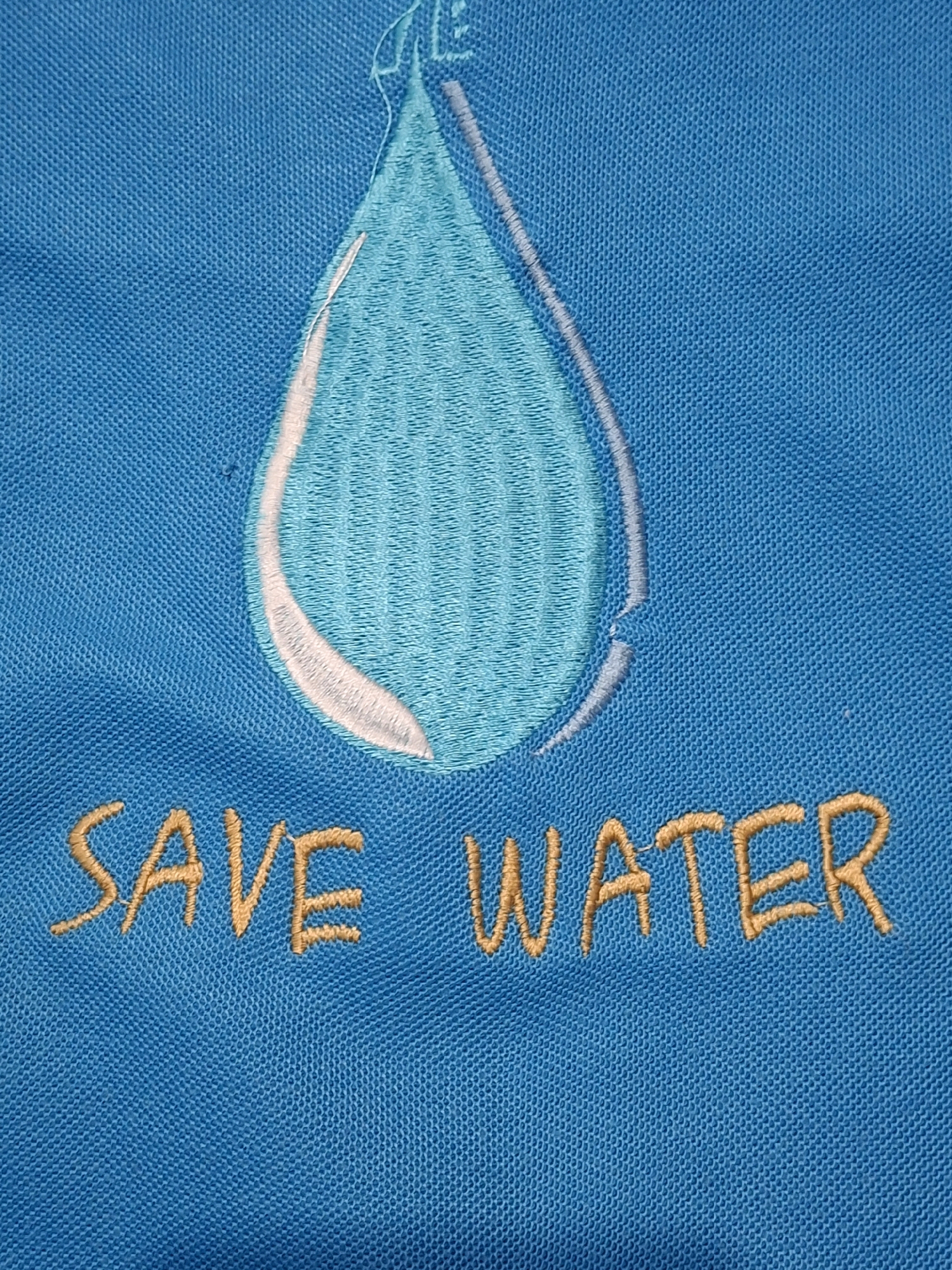A close-up, top-down shot shows embroidered text and a water droplet on a blue fabric. The text "SAVE WATER" is embroidered in a light brown thread across the bottom of the image. Above the text is a stylized water droplet, embroidered with various shades of blue and white thread, appearing to be a light blue drop with a darker blue interior and white highlights. The fabric has a visible woven texture. There is no discernible information to indicate the location in Lumajang, Indonesia.