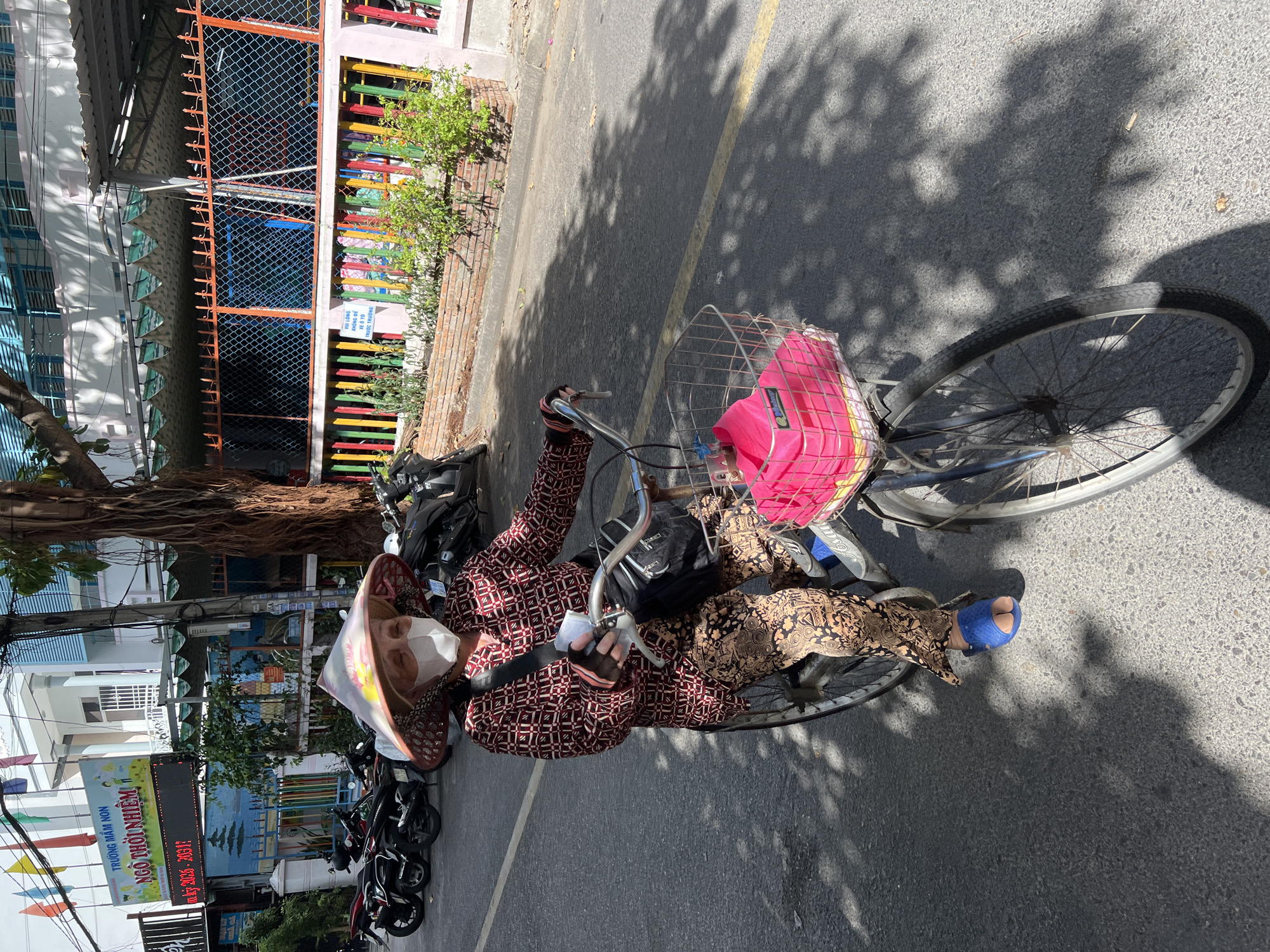 An elderly Vietnamese woman, wearing a conical hat and a face mask, rides a bicycle down a street in Nha Trang, Vietnam. She is dressed in a patterned maroon shirt and patterned brown pants. The bicycle has a metal basket on the front containing a bright pink bag. The street is paved with asphalt and has a yellow dividing line. In the background, there are several parked motorcycles, a building with a colorful fence, and trees. The scene appears to be in front of a school, indicated by signage on the building. The sunlight casts shadows on the street, suggesting it is daytime.