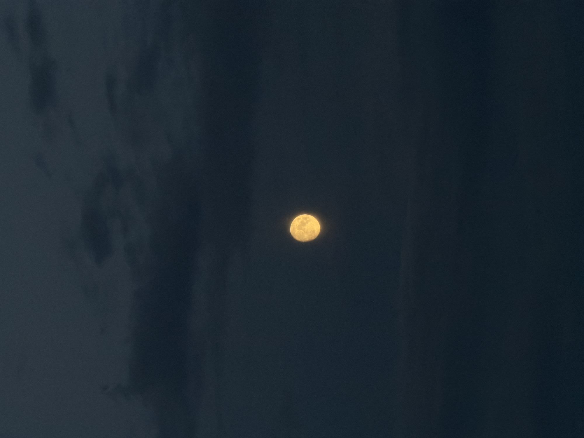 The image shows a full moon against a dark, cloudy sky. The moon is bright yellow and appears in the center of the frame. The clouds are dark and wispy, obscuring parts of the sky. The scene is dark, suggesting it is nighttime. There are no people, structures, or any indication of a specific location within the city of Khimki, Russia.