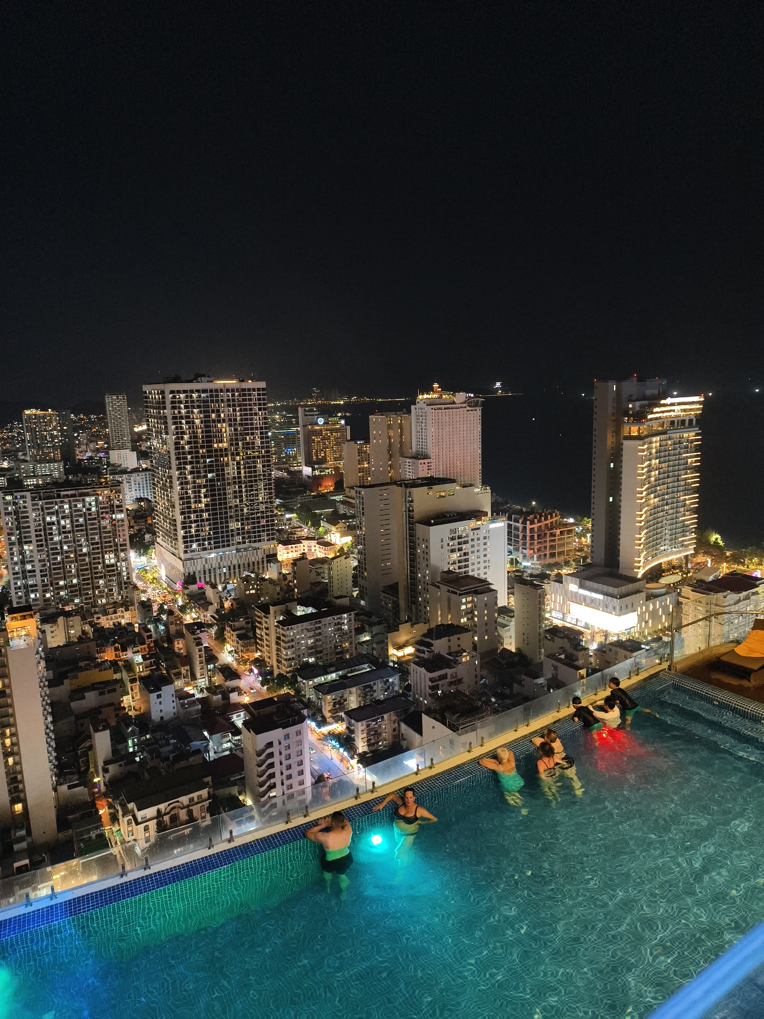 This high-angle night shot captures a vibrant cityscape from a rooftop perspective, featuring an infinity pool in the foreground. Several people are relaxing or swimming in the illuminated pool. To the left, a person in a dark swimsuit is partially submerged, glowing with a blue-green light from beneath the water. Another individual in dark swim trunks leans against the pool edge on the right. Further in the pool, a group of four people, including two women and two individuals in darker swim attire, are gathered, illuminated by red and green underwater lights.
The pool itself features blue and green tiles, and a clear glass barrier provides an unobstructed view of the urban expanse beyond. The city is densely packed with numerous high-rise buildings of varying designs and heights, all brightly lit from within their windows and by streetlights below. These lights create a complex network of glowing patterns against the dark night. In the distance, a vast body of water, likely an ocean or sea, is visible, with faint lights dotting its surface, suggesting distant ships or landmasses. The sky overhead is a deep, uniform black, indicating a clear night. The scene conveys a sense of relaxation and enjoyment, with the people appreciating the impressive nocturnal city vista. No visible text is present.