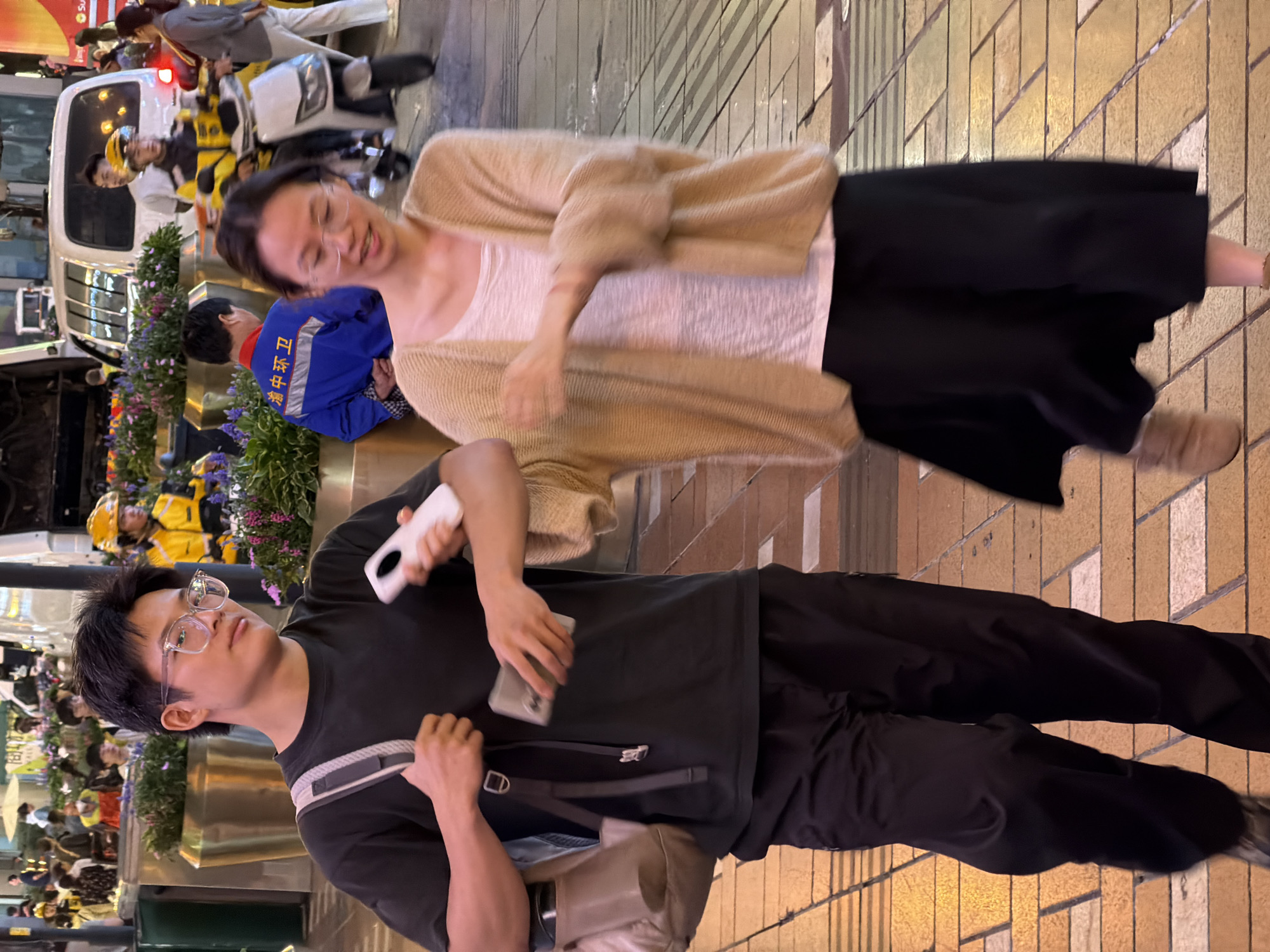 In Yuzhong District, China, two individuals are walking on a street at night. The person on the left is a young male wearing a black t-shirt and black pants, carrying a backpack and holding a phone. He has short dark hair and is wearing glasses. The person on the right is a young female wearing a light-colored cardigan over a white top and a black skirt. She is also wearing glasses and appears to be in motion. In the background, there are delivery riders on motorcycles, a white van, and a crowd of people. The street is paved with yellow and brown tiles, and there are planters with purple and pink flowers lining the walkway. The scene is illuminated by streetlights and the lights from surrounding buildings.