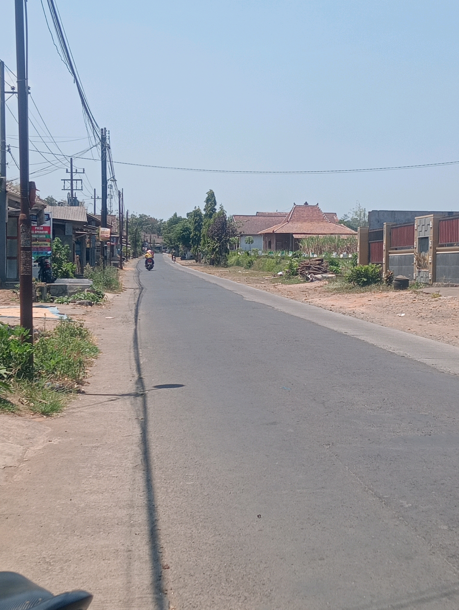 The image depicts a sunlit, rural street in Jatisari, Indonesia. The road is paved and appears to be two lanes wide, with a light gray asphalt surface. Vegetation, including grass and some small trees, lines the sides of the road. On the left side of the road, there is a sidewalk made of concrete. Several utility poles with numerous wires stretch across the scene, indicating infrastructure. Buildings are visible along the street, mostly single-story structures with tile roofs. Some of these appear to be houses, while others might be small shops or businesses, as suggested by signage on one of the buildings. In the distance, a motorcycle with two people on it is traveling away from the camera. The sky is clear and blue, suggesting a warm, dry day. The overall impression is of a quiet, everyday scene in a village setting.