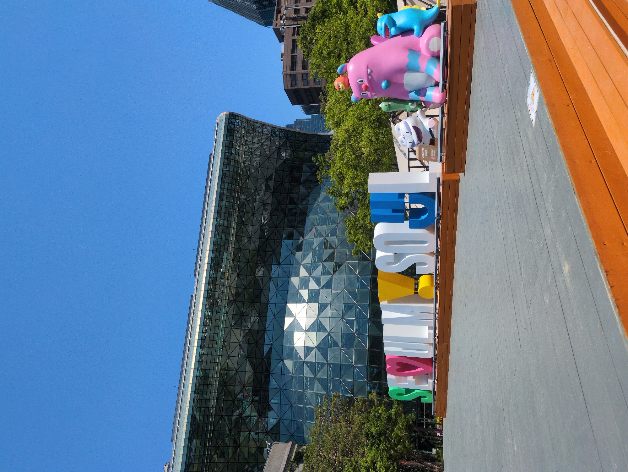 The image shows a partial view of the Coex Aquarium sign in Seoul, South Korea. The sign reads "SEOUL SUMMER VACATION" in colorful block letters. A building with a reflective triangular-patterned facade stands adjacent to the sign, partially obscured by trees. The image also features a wooden boardwalk and a blue sculpture of cartoon-like animals atop the sign. The sky is clear and blue. The scene appears to be taken during daylight.