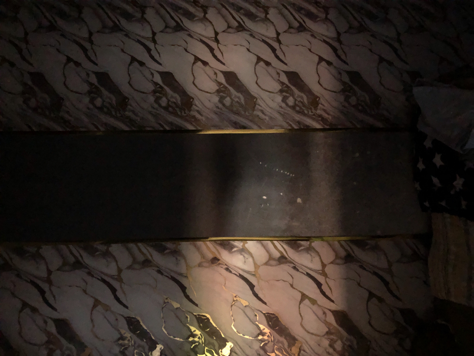 The image shows a surface with a marble-like pattern in shades of gray, white, and brown with dark veining. A dark gray, possibly metallic, rectangular object is positioned horizontally in the center of the frame, appearing to be bordered by gold trim. In the top right corner, a portion of a pillow or cushion with a star pattern is visible. The lighting is dim, casting shadows across the scene. The image may be in Rantopanjang, Indonesia, but there are no elements in the picture that can confirm the city as the source of the picture.