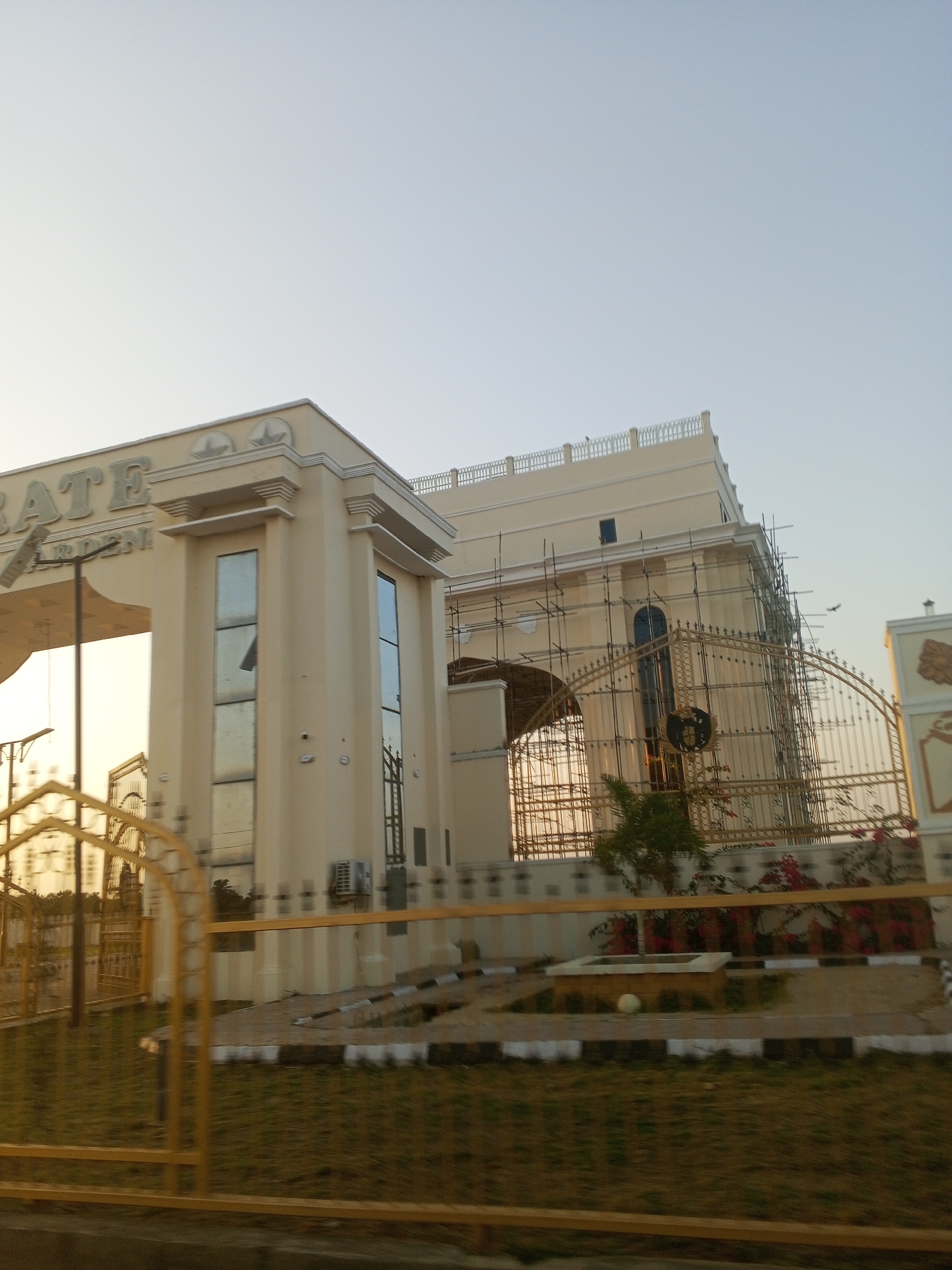 The image shows a partial view of an entrance to what appears to be a property in Tawpo, Nigeria. The entrance features a large, off-white gate with the word "GATE" and "GARDEN" visible on the structure. To the right of the gate, a building is under construction or renovation, with scaffolding visible around its façade. A decorative golden metal fence surrounds the property. A small, square planter sits in front of the fence, containing a white ball object. Green grass and some plants are visible within the enclosed area. The lighting suggests it is either early morning or late afternoon. The sky is clear and pale.