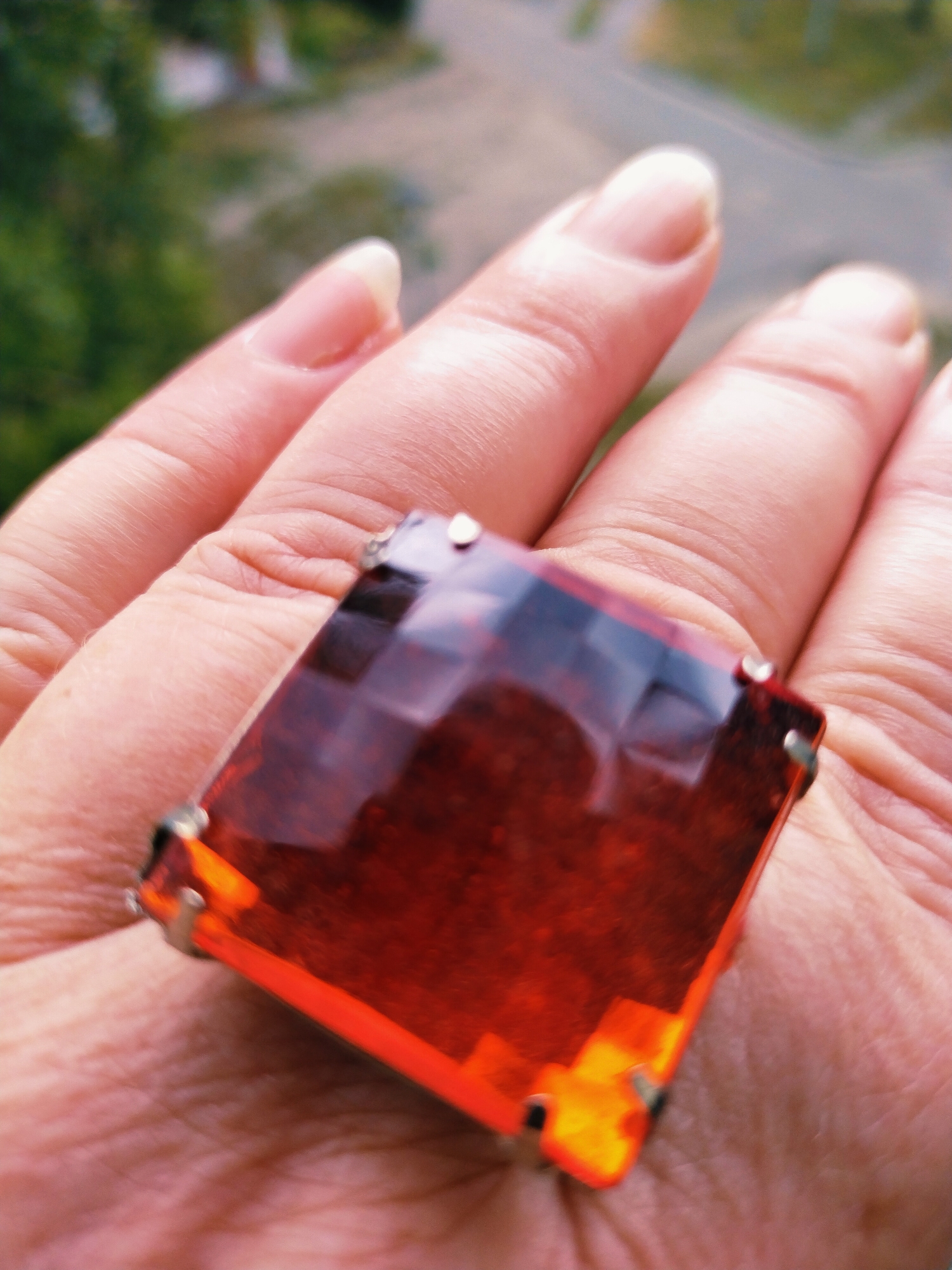 A close-up shot shows a person's left hand, with fingers extended, wearing a large, square-shaped amber ring. The ring features a faceted, translucent amber stone set in a silver-toned metal. The stone is a deep reddish-brown color with internal reflections and variations in hue. The hand exhibits visible skin texture and wrinkles. The background is blurred, with hints of green foliage and a light-colored surface, possibly a road or pavement, suggesting an outdoor setting in Horishni Plavni, Ukraine. The lighting appears natural, casting subtle shadows on the hand and highlighting the facets of the gemstone.