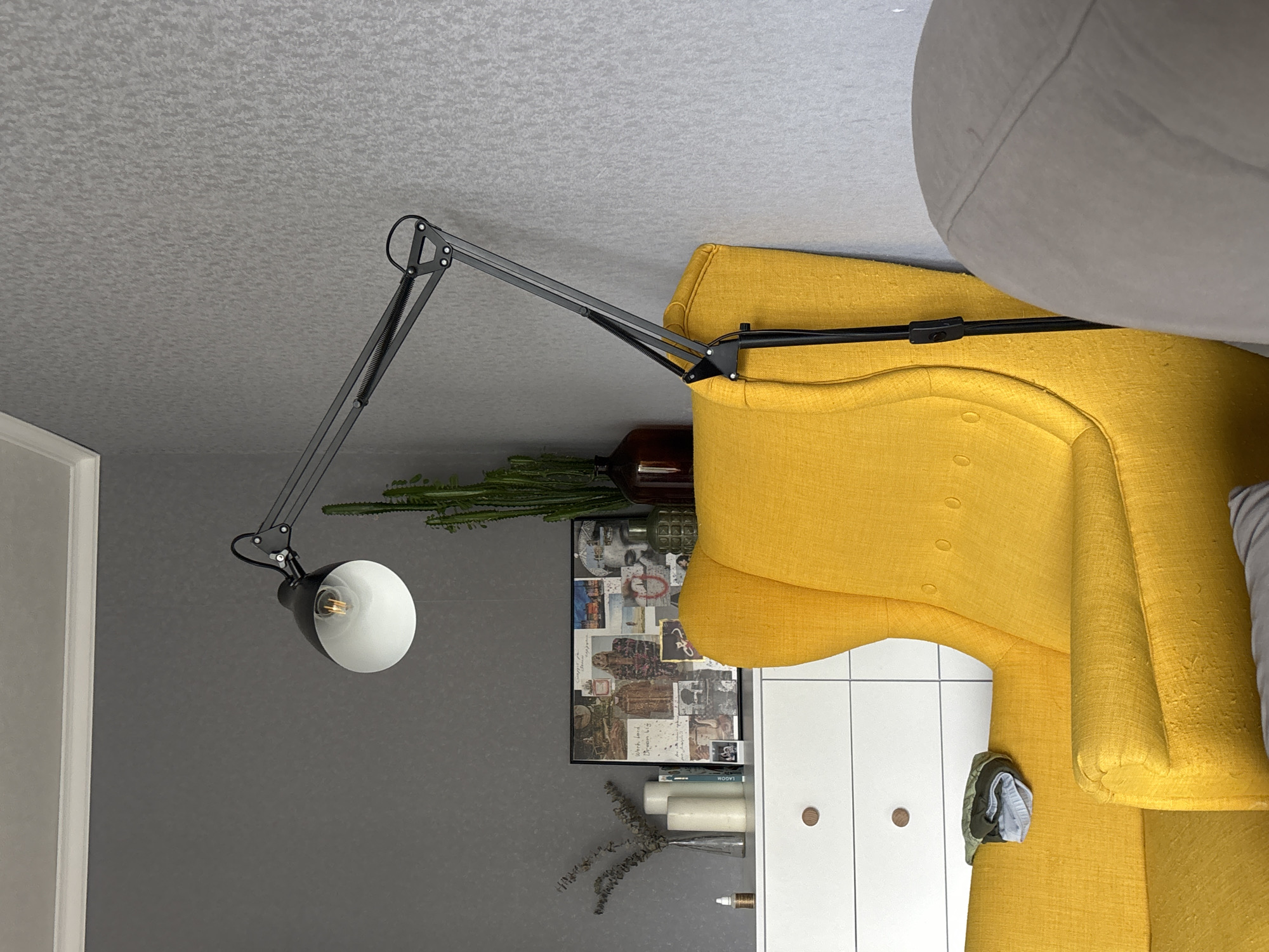 A yellow armchair is adjacent to a white cabinet, with a black desk lamp positioned over both. The lamp is illuminating the area. A framed collage is on the wall behind the cabinet, along with some plants and decorative objects. The armchair is partially obscured by a grey sofa. The room has grey walls and a textured ceiling. The location is a room, and there is no information indicating this is Chaikovsky, Russia.