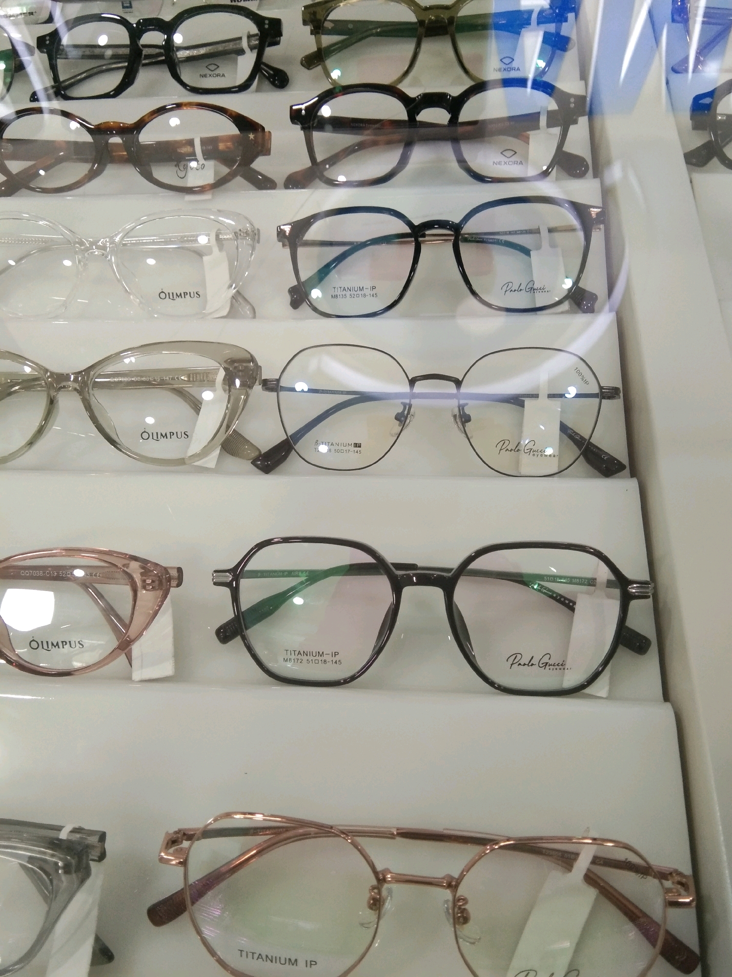 The image shows a display of eyeglasses in a retail setting. The eyeglasses are arranged on white shelves, and some have labels indicating brand names such as "NEXORA," "OLUMPUS," and "Paolo Gucci Eyewear." The lighting is bright, suggesting an indoor environment, possibly a shop or a market stall. There are no people visible in the image. The overall scene is clean and organized, with the focus on the merchandise. The city of Kebumen, Indonesia, is not depicted or referenced in the image.