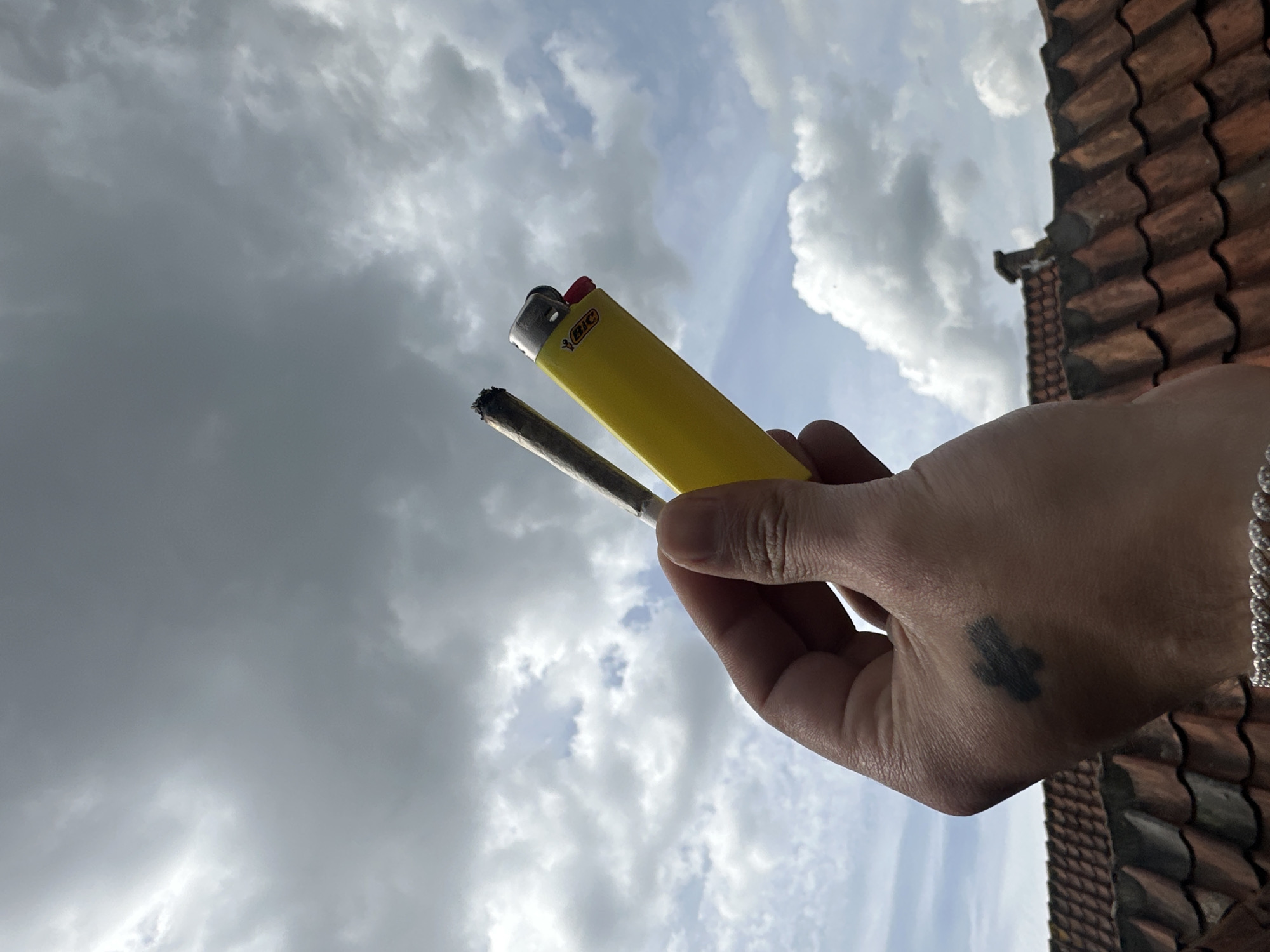 The image, captured in Amersfoort, Netherlands, is a low-angle shot looking upwards towards a dynamic, cloudy sky. In the foreground, a human hand holds a bright yellow BIC lighter and a hand-rolled cigarette, commonly referred to as a joint. The tip of the cigarette appears partially burned, suggesting it is either lit or has recently been used.
The hand, seen from the back, features a dark, abstract or cross-like tattoo on its surface and a silver chain bracelet on the wrist. The "BIC" logo is clearly visible on the yellow lighter. The background consists predominantly of a partly cloudy sky, with a mix of dark grey, heavy clouds and brighter white clouds, indicating a daytime setting. Some patches of lighter blue sky are visible between the clouds. On the right side of the frame, a section of an old, reddish-brown tiled roof is visible, providing a contextual clue for an outdoor, elevated location such as a balcony or rooftop.