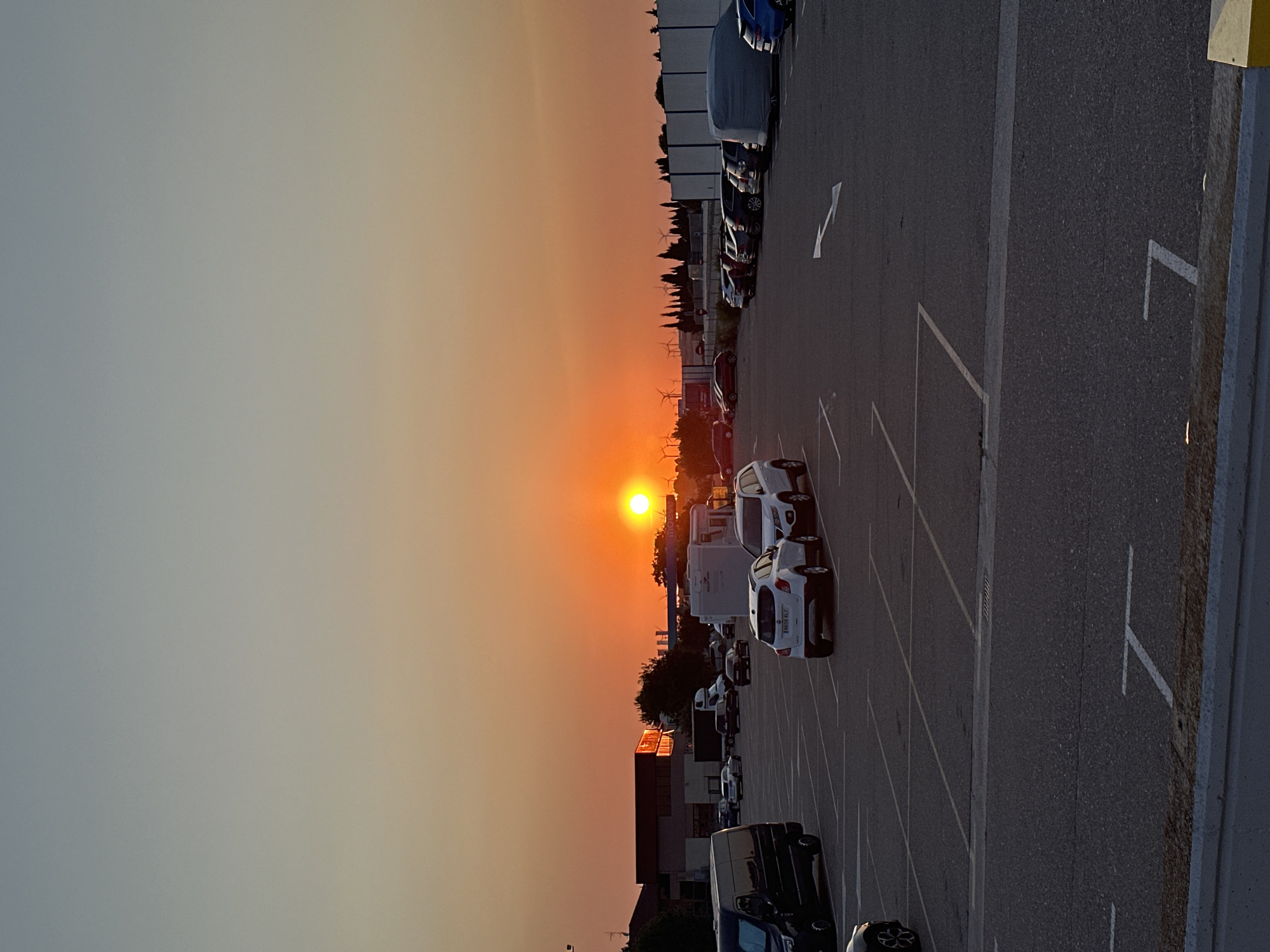 An outdoor, eye-level, full shot captures a parking lot at sunset in Pedrola, Spain. The sun, a bright orange orb, is positioned in the center of the frame, near the horizon, casting a warm glow across the sky and a vast, empty parking lot. The sky transitions from a pale blue at the top to a vibrant orange, tinged with pink, towards the horizon.
The parking lot, paved with asphalt and marked with white lines, is populated by several parked cars. A black van is parked on the left side of the frame, partially obscuring a white car. In the middle ground, a white SUV is parked in a designated spot, facing away from the camera, with another white SUV parked to its right. Further back, a row of cars is visible, including a blue car on the far right. In the distance, buildings, including what appears to be a commercial establishment with a red roof, and a gas station are visible. A line of trees and windmills can be seen against the orange sky. The foreground of the parking lot shows clear lane markings. The overall impression is one of a serene, end-of-day scene.