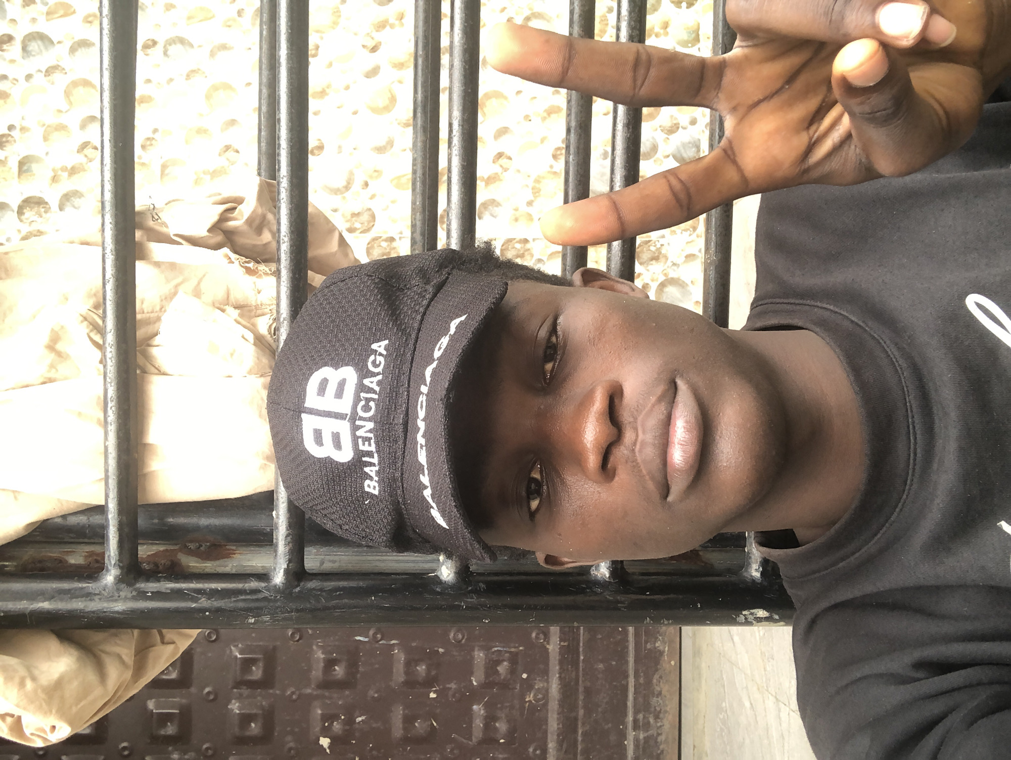A young Black male with dark skin, wearing a black Balenciaga baseball cap and a black crewneck sweatshirt, poses for a selfie in front of a metal gate. He has a neutral expression and is holding up his right hand in a peace sign. Behind him, a patterned wall is visible, with some brown fabric draped over the gate. The scene appears to be outdoors, possibly on a street or in a courtyard, given the metal gate and textured ground. The lighting is somewhat harsh, casting shadows on his face and hands. The overall setting suggests an urban environment, consistent with a location in Ado, Nigeria.