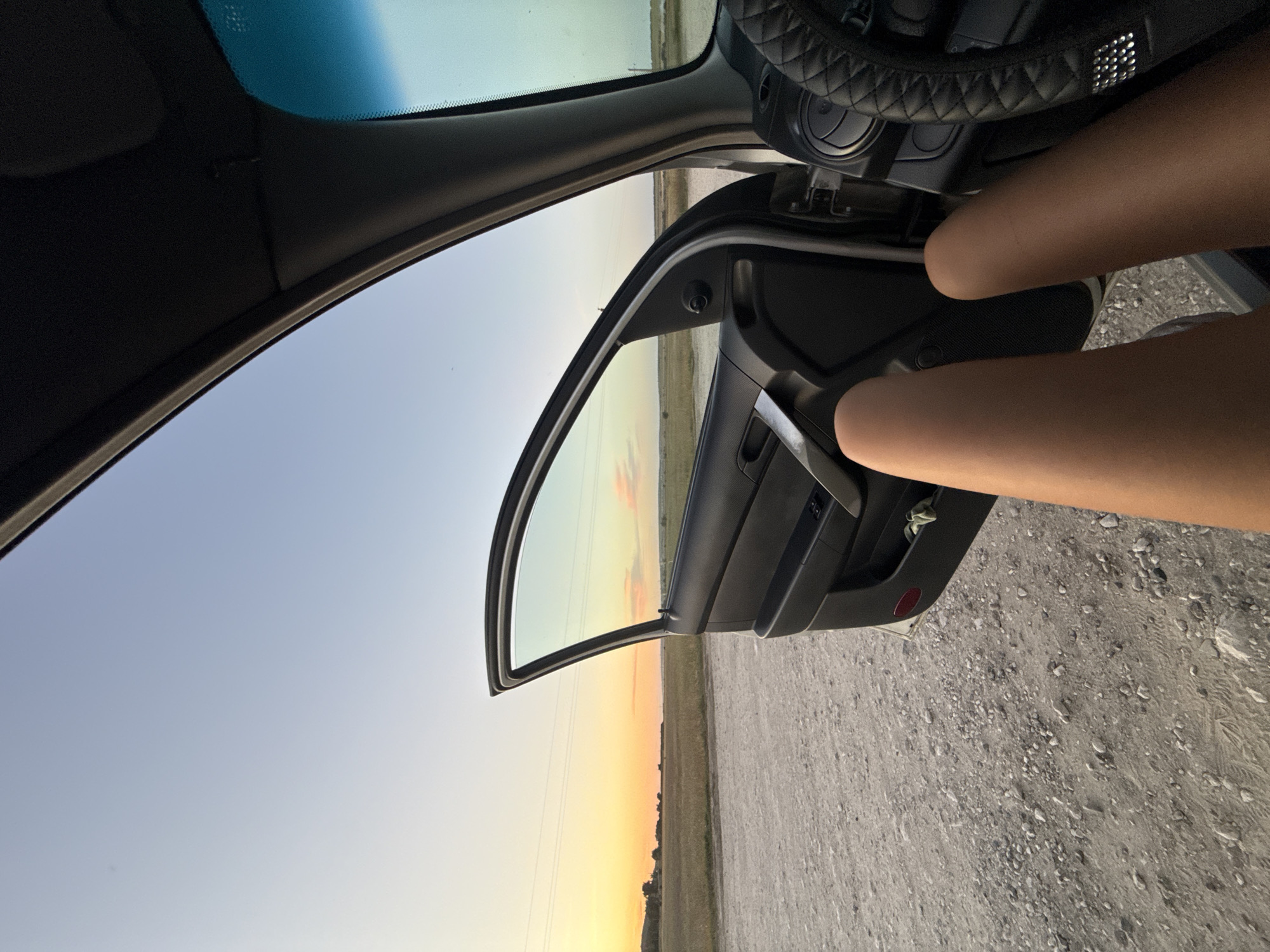 The image depicts the interior of a car parked on a gravel road. The driver's side door is open, revealing a rural landscape with a sunset in the background. A person's legs are visible, suggesting they are sitting inside the car. The sky is clear with hints of pink and orange from the sunset. The road is made of light-colored gravel and small stones. Power lines are visible in the distance. The car's interior shows the door panel, a steering wheel, and part of the dashboard. The scene appears to be in an unpopulated, open area.