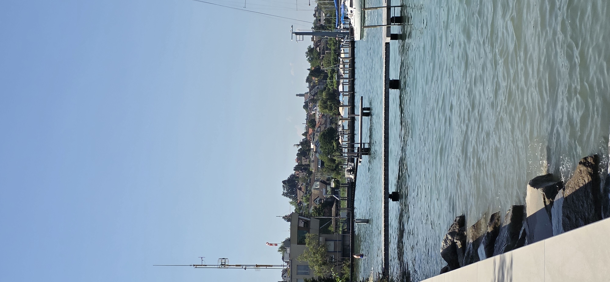 The image depicts a waterfront scene in Oberrieden, Switzerland, on a clear, sunny day. In the foreground, a concrete promenade runs along the left edge of the frame, bordering a rocky shoreline. The water, which appears to be a lake, is calm with small ripples. Several wooden piers extend into the water, with one prominent pier in the middle ground. A person is visible in the water near the left side of the frame, seemingly swimming or wading.
In the midground, a cluster of moored boats are visible, some with white covers. A white sailboat with a mast is docked on the right side. Behind the waterfront, a hillside town is visible, characterized by numerous houses with red-tiled roofs and lush green trees. A prominent telecommunications tower with an anemometer and a windsock stands on the left side of the town. The sky is a clear, pale blue. The overall scene conveys a peaceful, leisurely atmosphere typical of a summer day at a lakeside town.