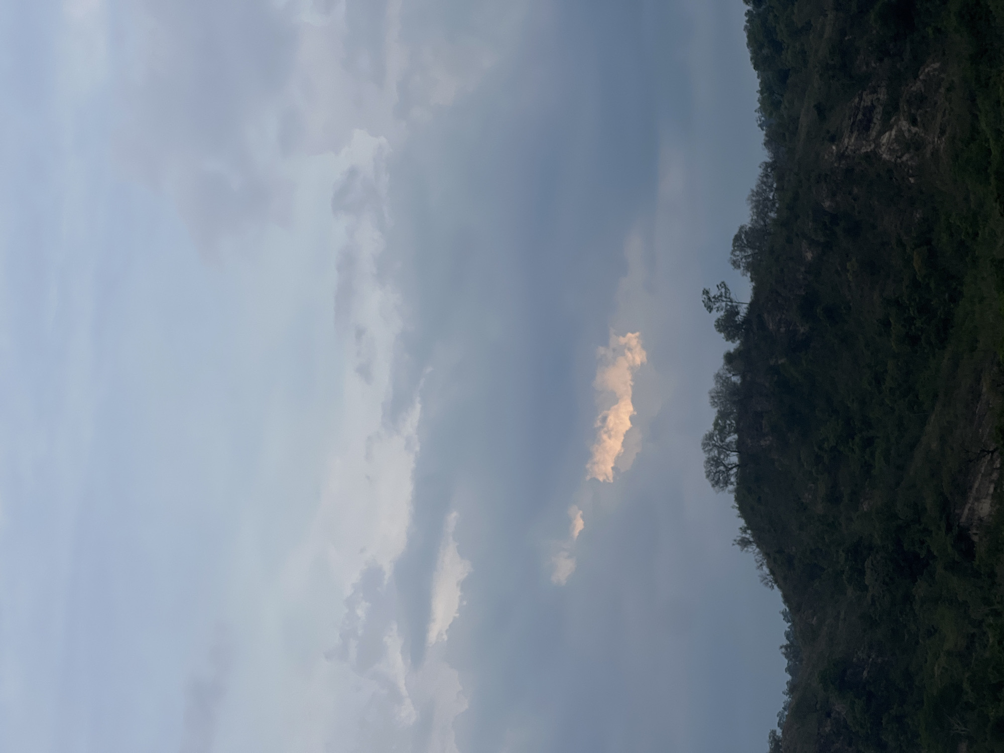 The image shows a view of the sky with clouds and the silhouette of a forested mountain ridge on the right. The sky is mostly overcast with varying shades of gray and some hints of light breaking through the clouds. The mountain ridge is dark green, with visible trees along the top. There is no indication of location in Gracias, Honduras or any people, subjects, objects, actions, or interactions.