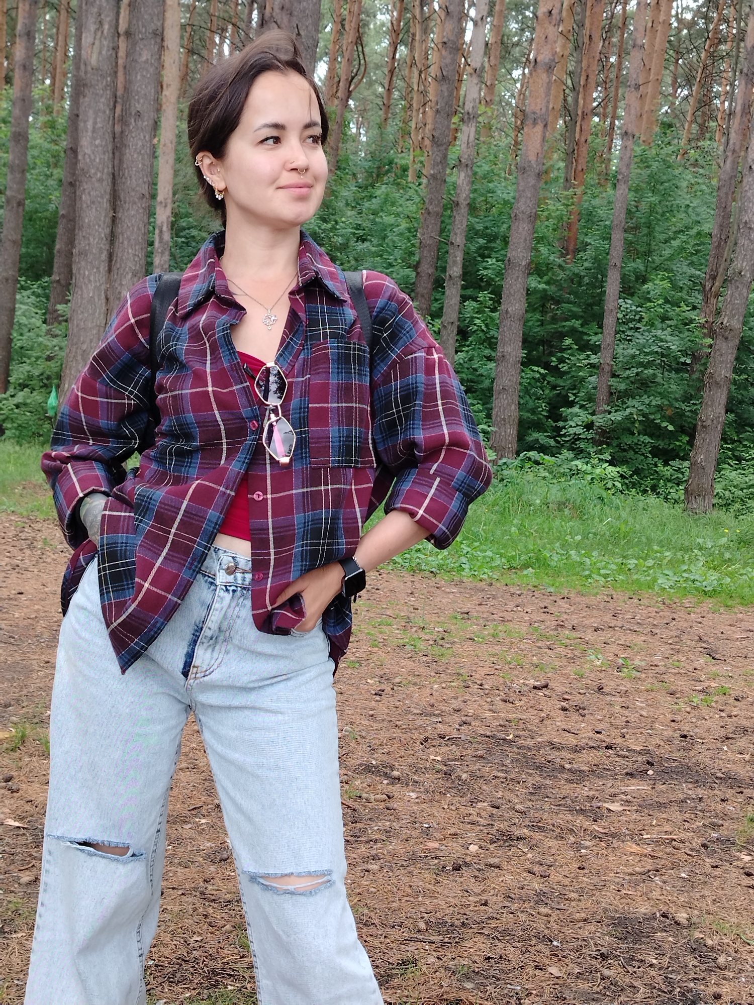 A young woman stands in a forest with pine trees and greenery. She has dark hair styled short, several ear piercings, and a nose ring. She is wearing a red tank top, an open, maroon and blue plaid shirt, light-wash ripped jeans, and a black wristwatch. She is accessorizing with a necklace and sunglasses hanging from her shirt. Her hands are on her hips, and she is looking off to the right. The ground is covered with pine needles and pine cones. The location is described as being in the city of Chaikovsky, Russia.