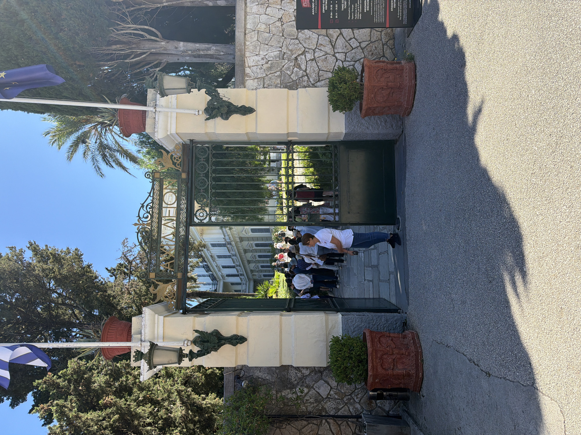 The image shows the sunny entrance to the Achilleion Palace in Gastouri, Greece, with a group of visitors gathered at the gate. An ornate, dark metal gate with decorative gold elements stands open, revealing a grand, multi-story white building with numerous windows, partially obscured by trees and lush greenery in the background.
On the left side of the entrance, Greek and European Union flags are visible, flying from flagpoles. A large potted palm tree and an antique-style street lamp further adorn this side. On the right, a stone and plaster pillar supports an information board. The ground in front of the gate is paved with a light-colored aggregate, with prominent shadows indicating bright daytime conditions and clear weather.
A group of people, mostly adults, are congregated inside the open gate, appearing to be queuing or waiting to enter the palace grounds. One person in a white t-shirt and blue jeans stands out in the foreground, facing into the estate.
Visible text on the information board confirms the location and provides visitor information. It reads "ΧΙΛΛΕΙΟ Α" (Achilleion), "ΩΡΑΡΙΟ ΛΕ VISITING" (Visiting Hours) with seasonal date ranges ("01.04 - 31.10" and "01.11 - 31.03"), "ΤΙΜΕΣ ΕΙΣ TICKET" (Ticket Prices) for "Groups," and "ΜΕΙΩΜΕΝ DISCOUN" (Reduced/Discount). The presence of the flags and the explicit mention of "Achilleion" on the sign firmly place the scene at the historical Achilleion Palace.